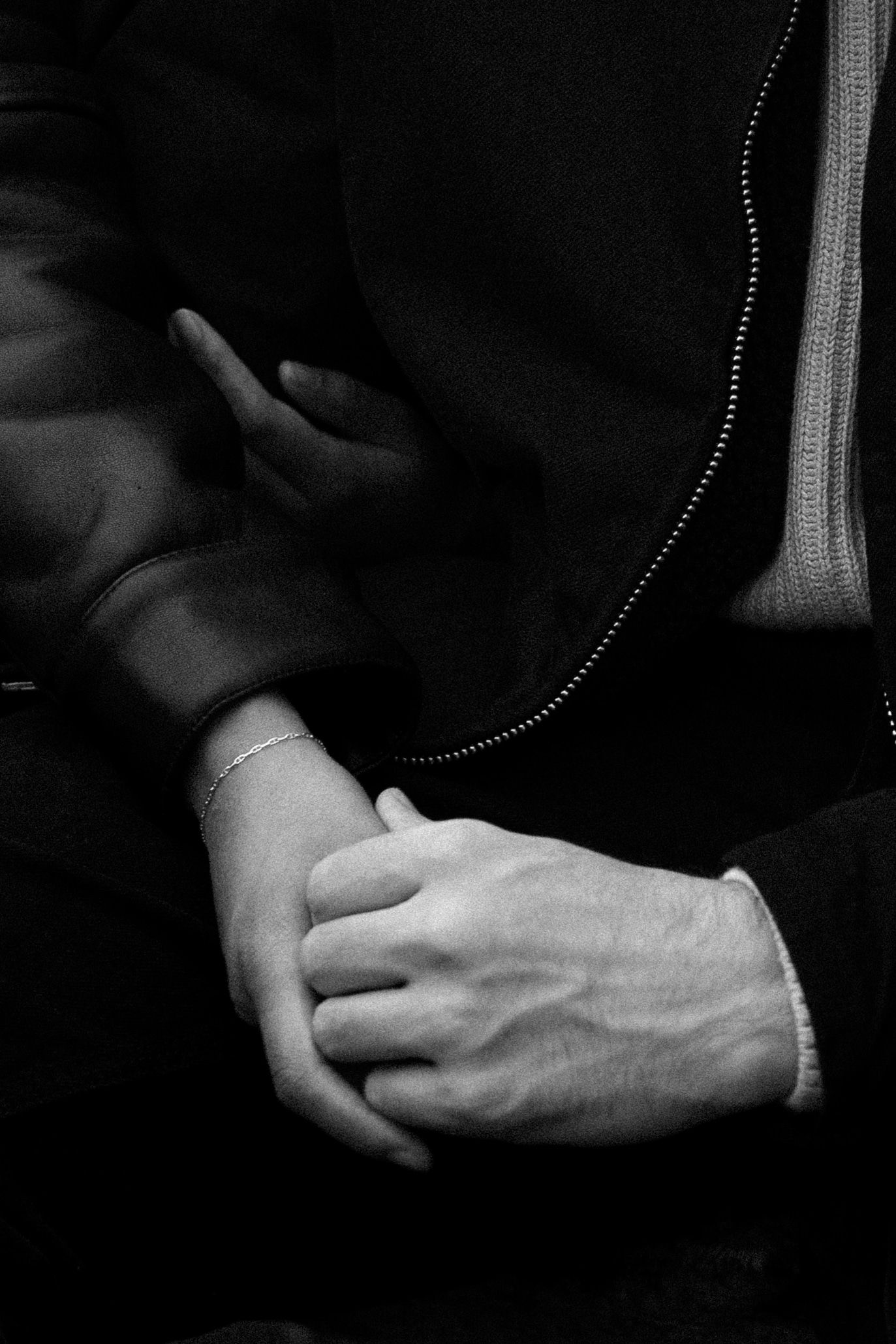 Black and white photo of two people holding hands, one wearing a delicate bracelet, dressed in dark clothing.