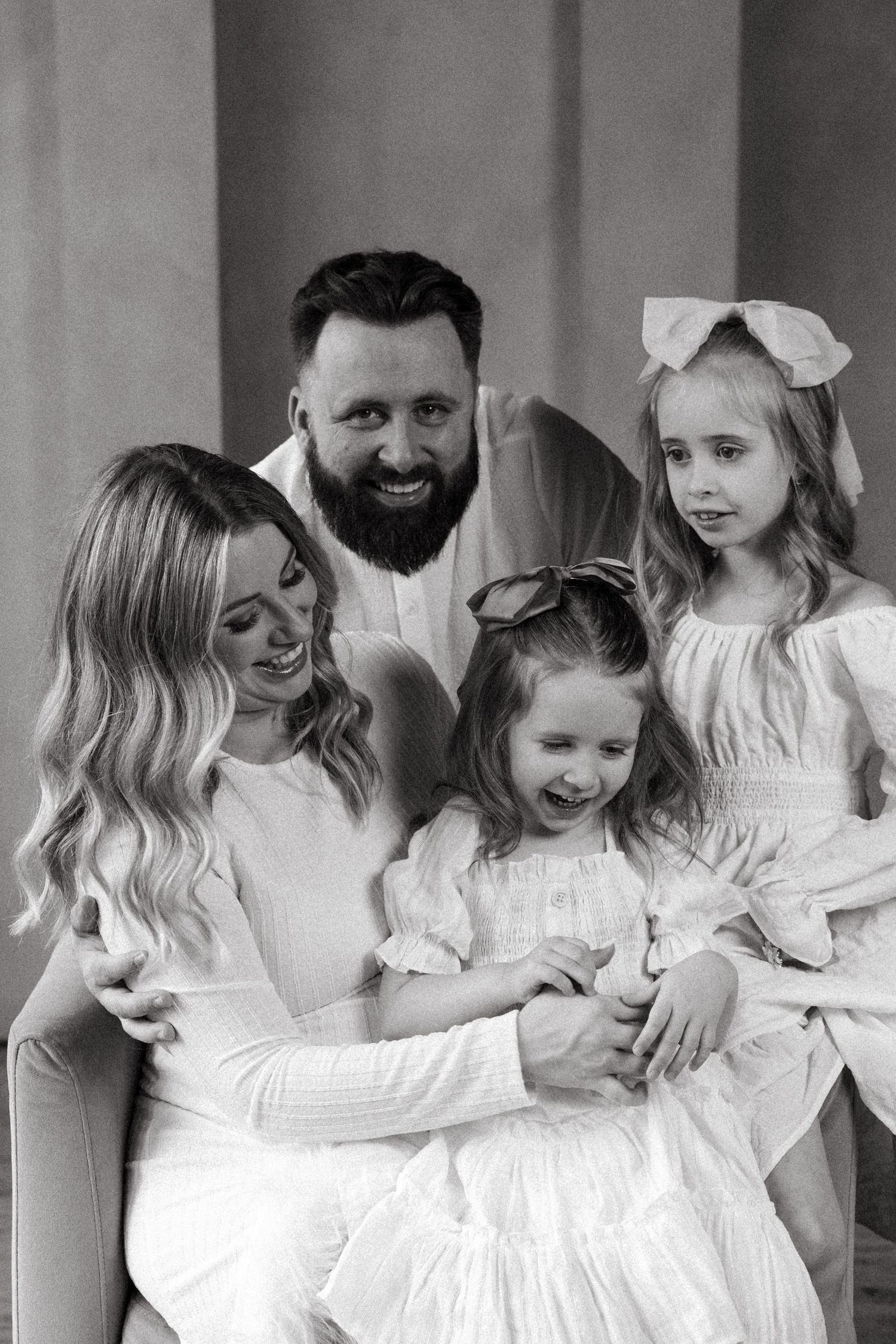 Black and white family portrait of smiling bearded man, blonde woman, and two young girls in white dresses with bows.