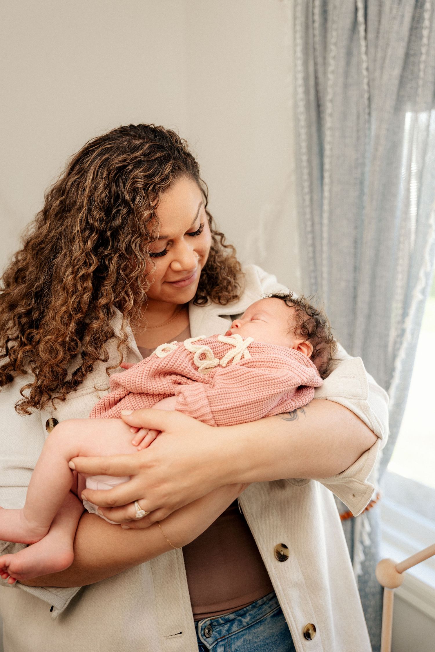A curly-haired woman lovingly cradles her newborn baby dressed in a pink outfit near a curtained window.