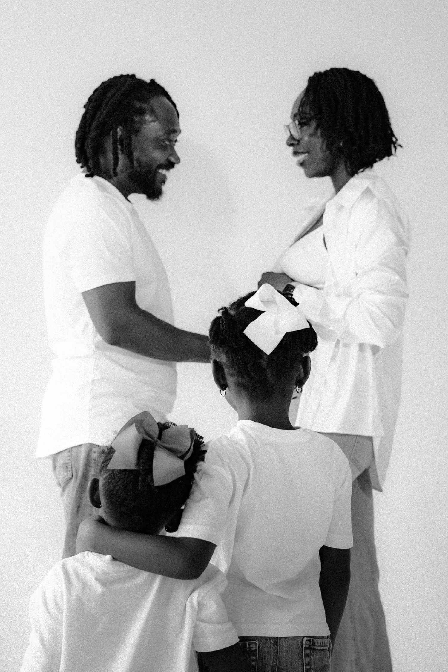 Black and white family maternity photo with parents in white outfits and two children hugging mother's pregnant belly.