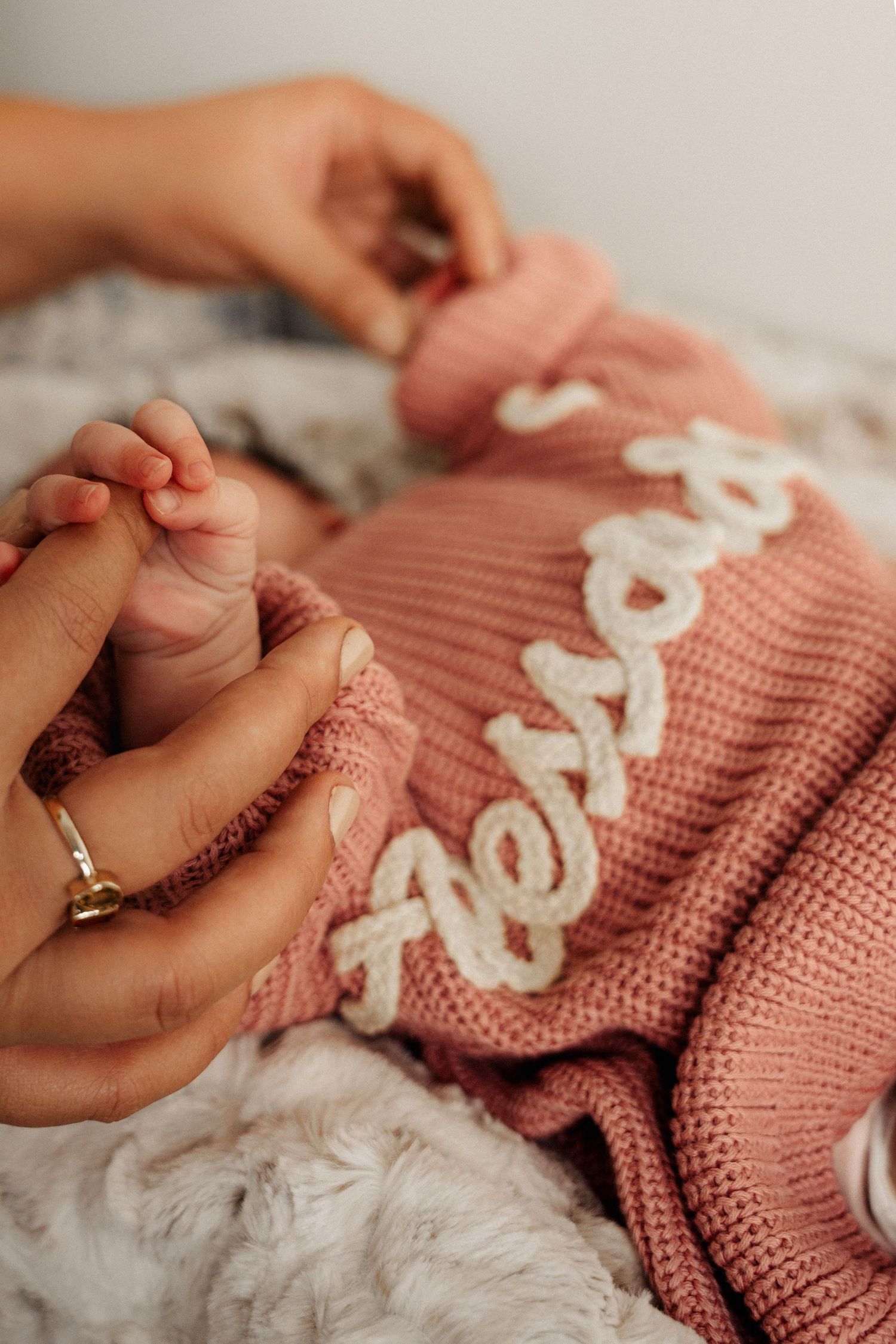 Newborn baby in pink knit sweater with name embroidery, gently holding mother's hand with gold ring.
