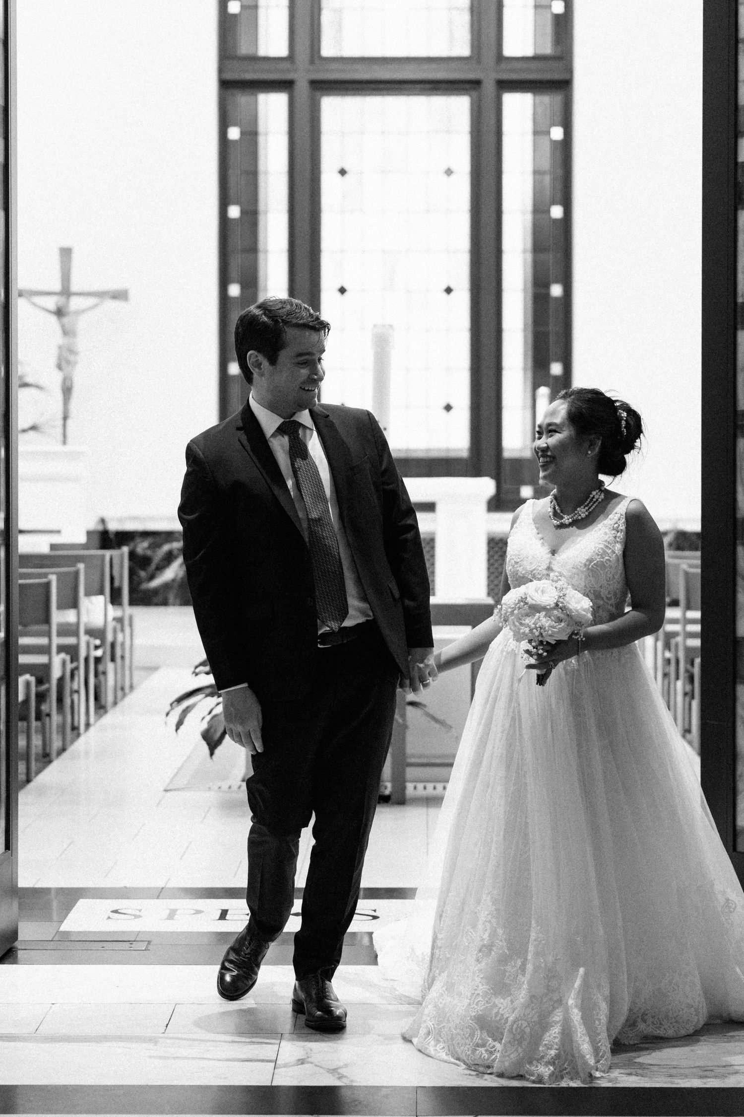 Black and white photo of a bride and groom walking hand in hand down a church aisle after their wedding ceremony.