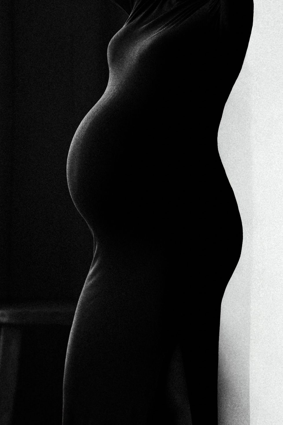 Artistic black and white silhouette profile of a pregnant woman in a fitted dress by a wall.