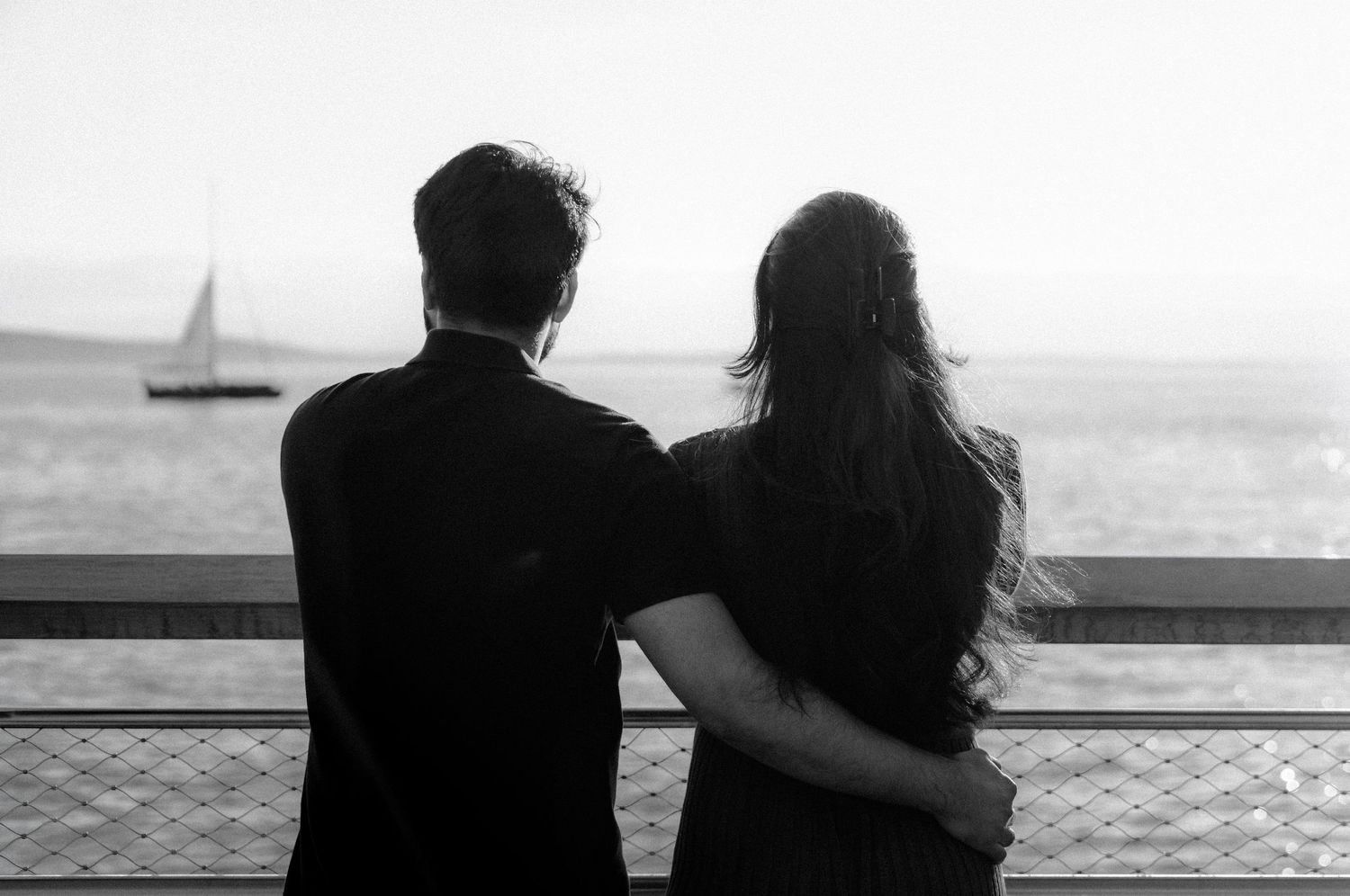 Couple embracing while gazing at a sailboat on the ocean from a waterfront railing in black and white.