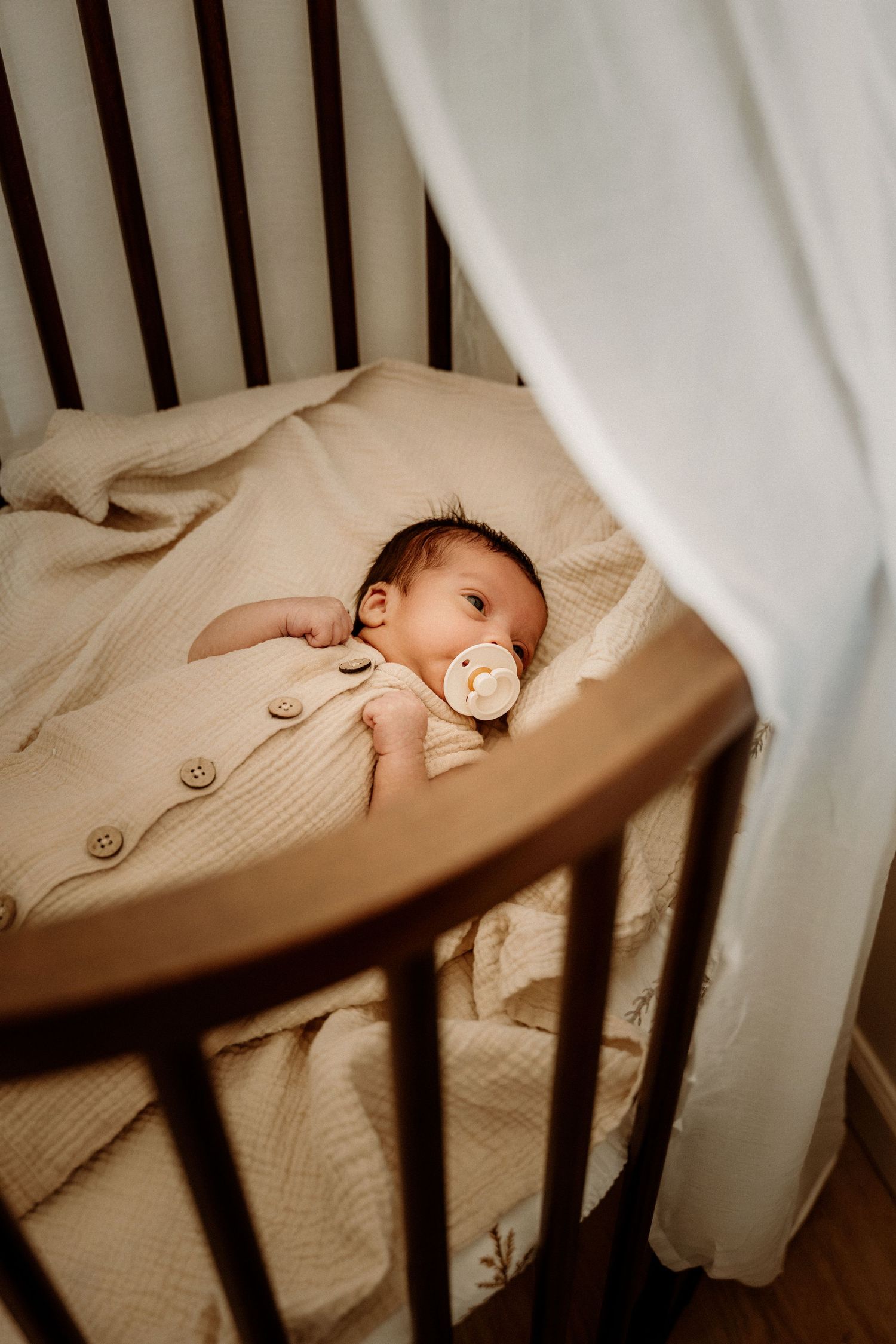 Newborn baby with pacifier lying in a dark wood crib with beige muslin blanket and white curtain.