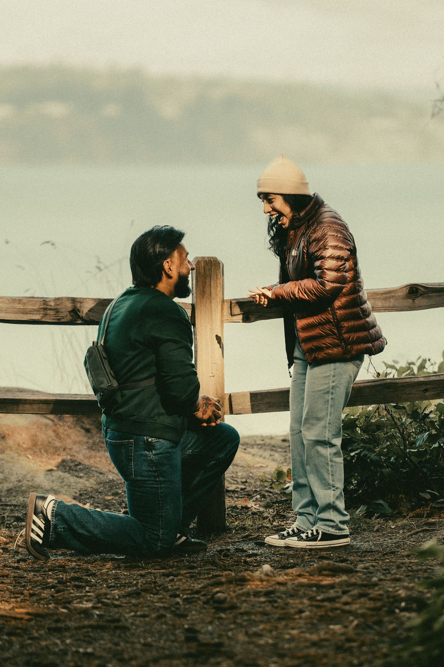 Man proposing on one knee to a smiling woman near a wooden fence outdoors on a cloudy day.