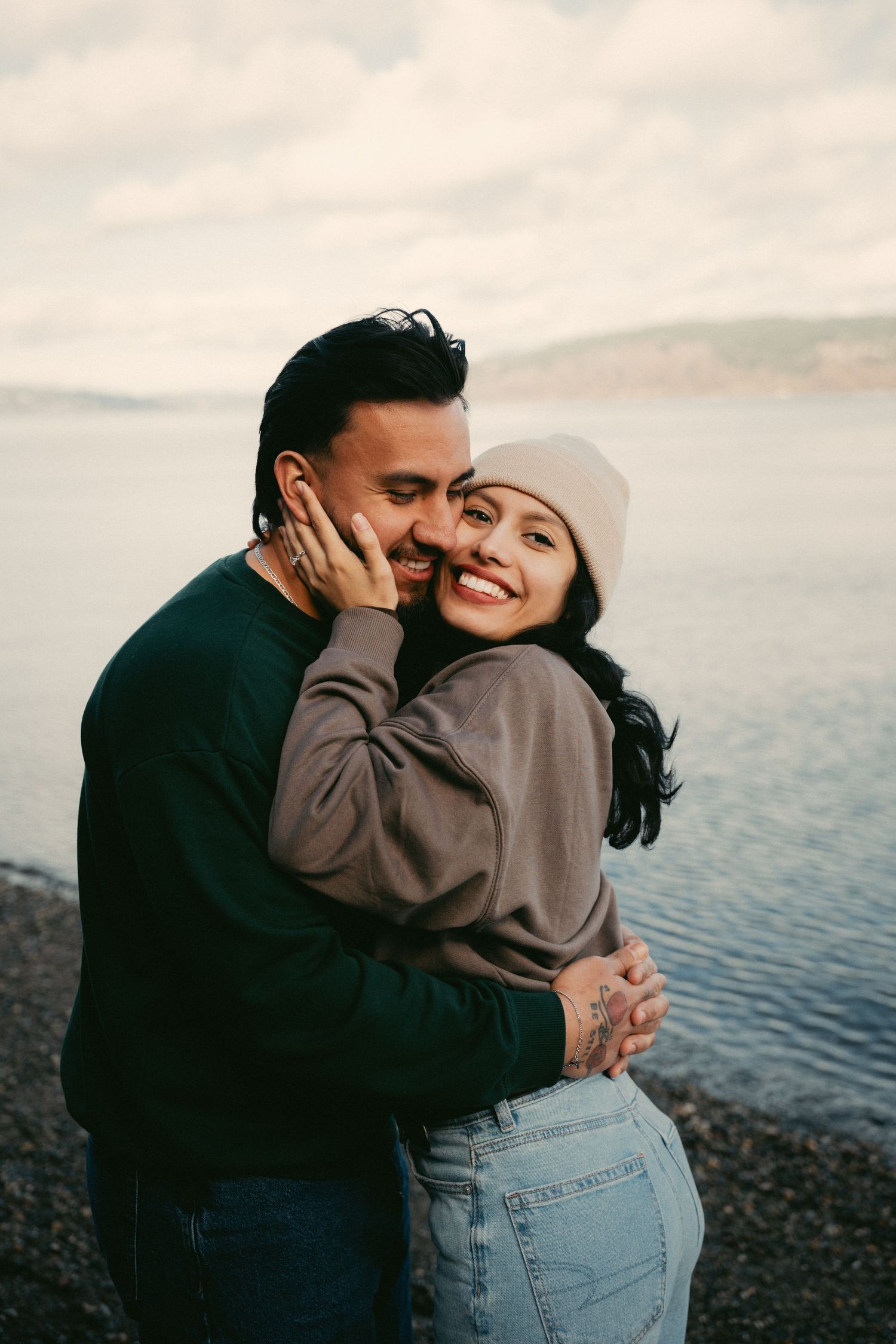 A couple embraces and smiles by a rocky beach with calm water and cloudy skies behind them.