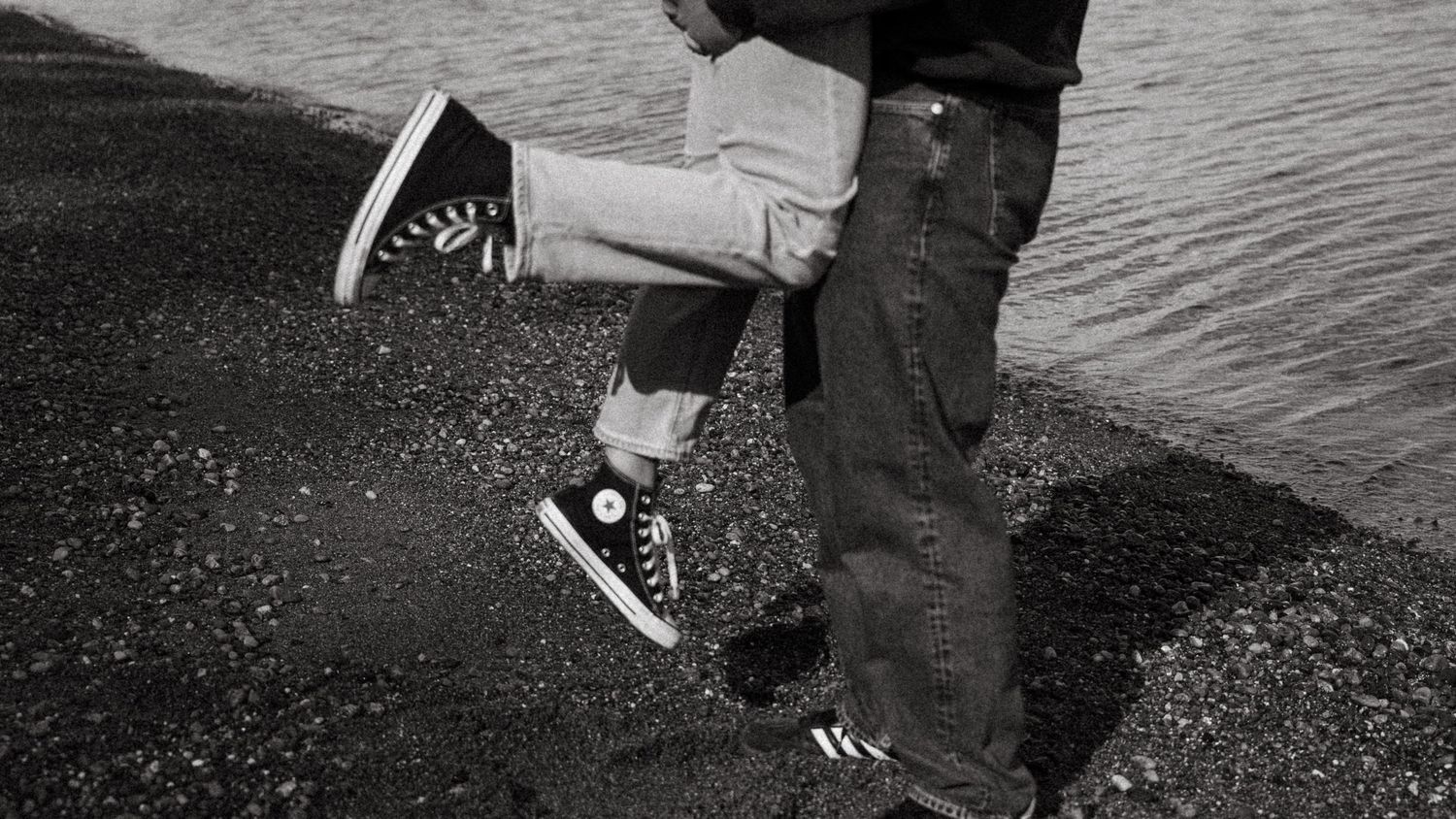 Black and white photo of a couple kissing, wearing Converse sneakers, standing on rocky ground near water.