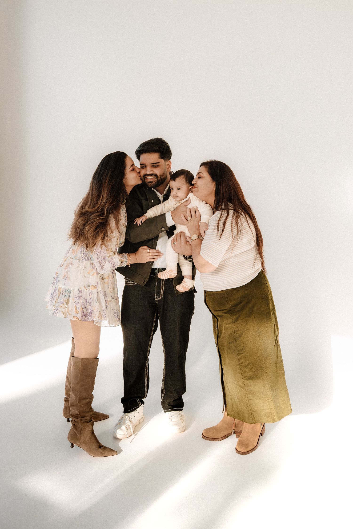Family portrait with man holding baby while two women kiss his cheeks in a bright white studio setting.
