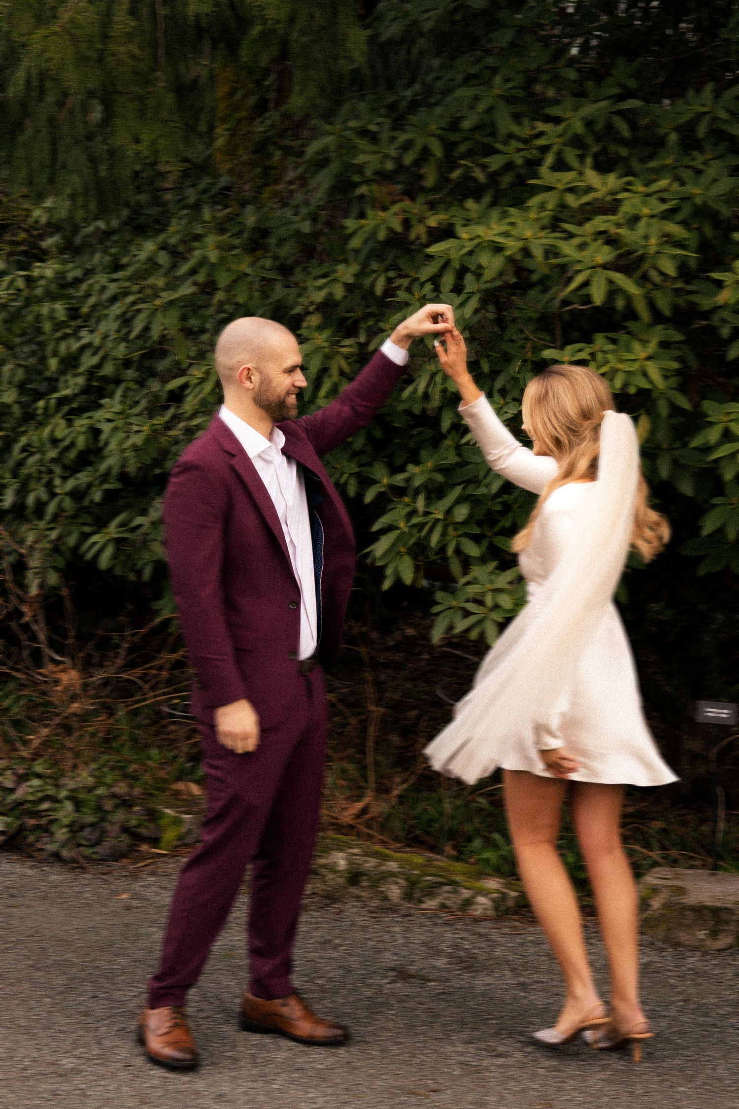 A bald groom in a burgundy suit twirls his blonde bride in a white mini dress outdoors.