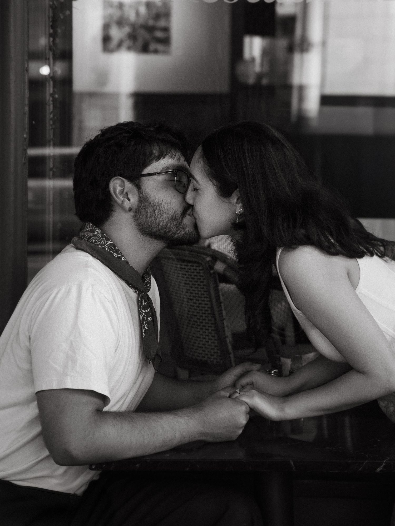 Black and white photo of a couple sharing a romantic kiss while holding hands at a cafe table.