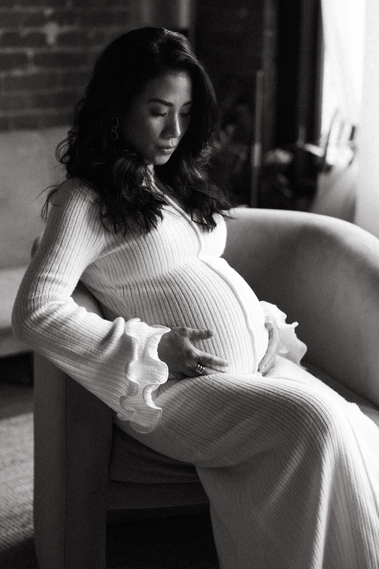Black and white photo of a pregnant woman in a ribbed white dress, sitting in a chair cradling her baby bump.