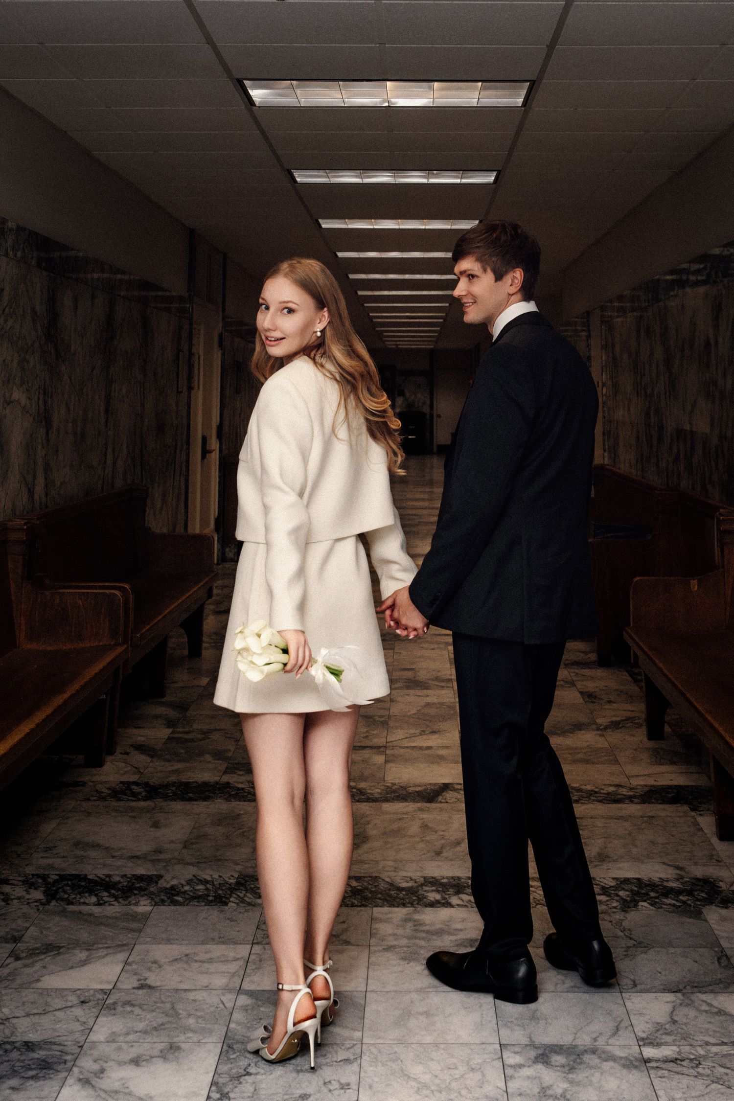 Elegant couple in wedding attire holding hands in a marble hallway, bride in white mini dress with roses.