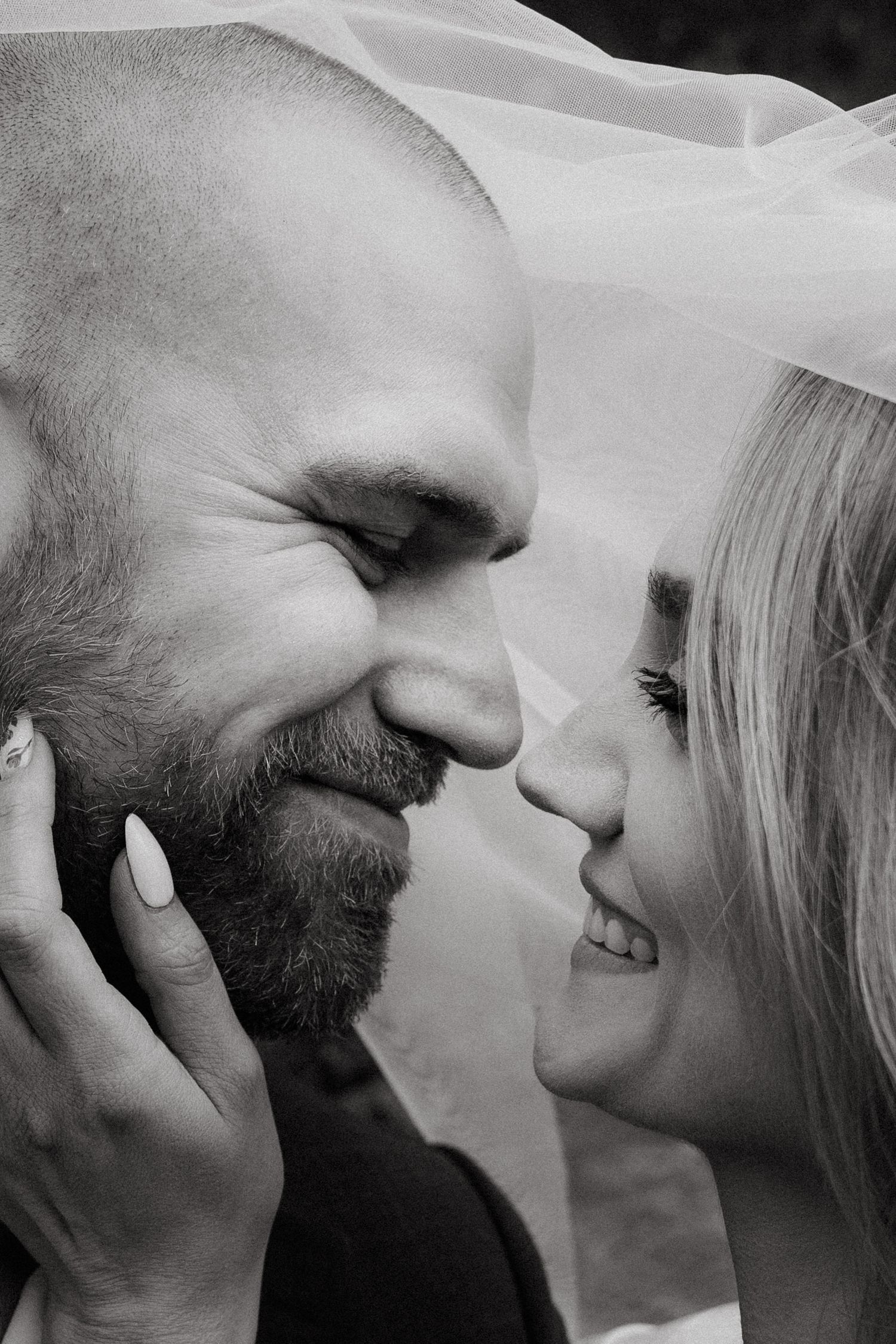 Black and white close-up of a smiling bride and bearded groom nearly kissing under a wedding veil.