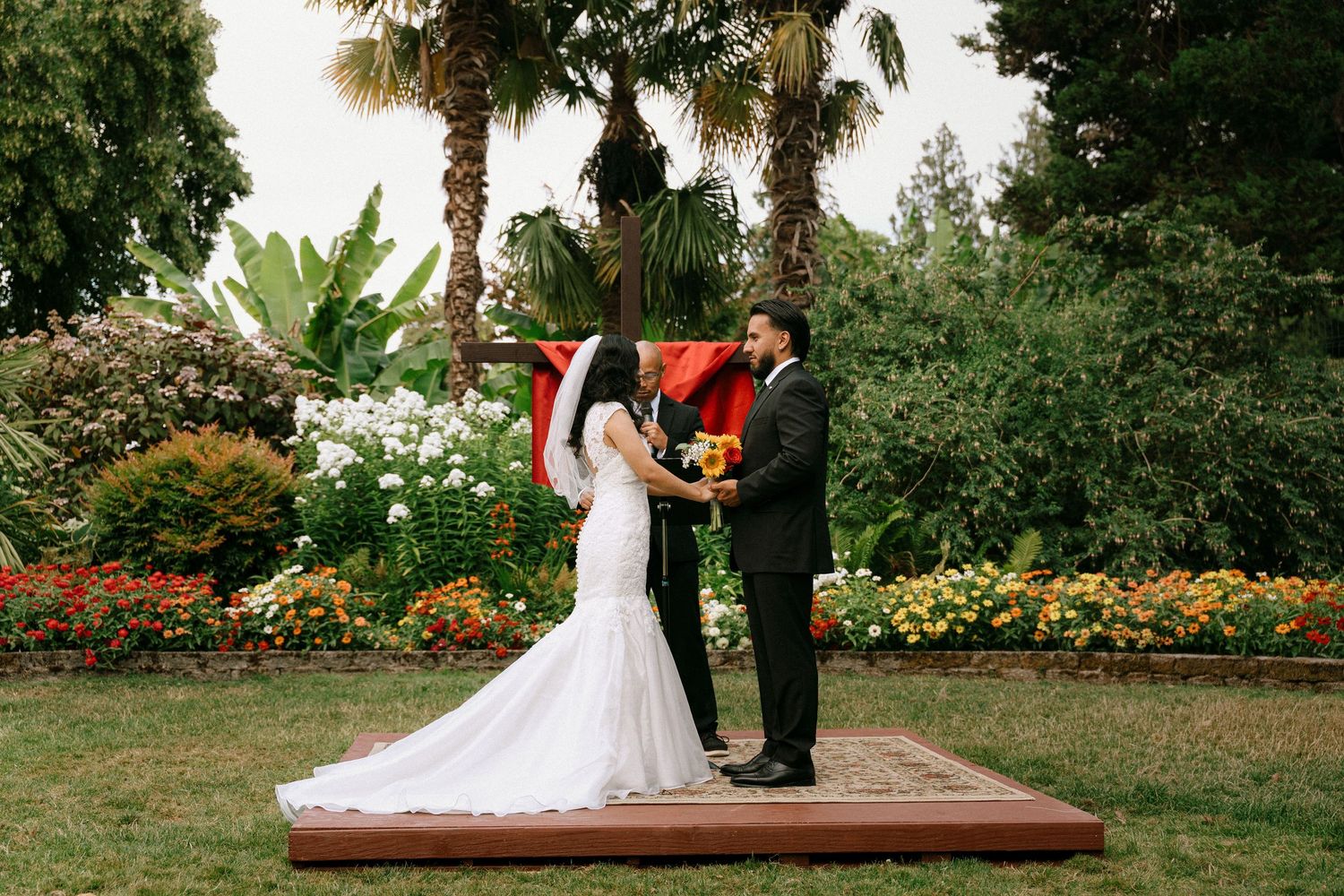 Bride and groom exchange vows outdoors on wooden platform surrounded by colorful garden flowers and palm trees.