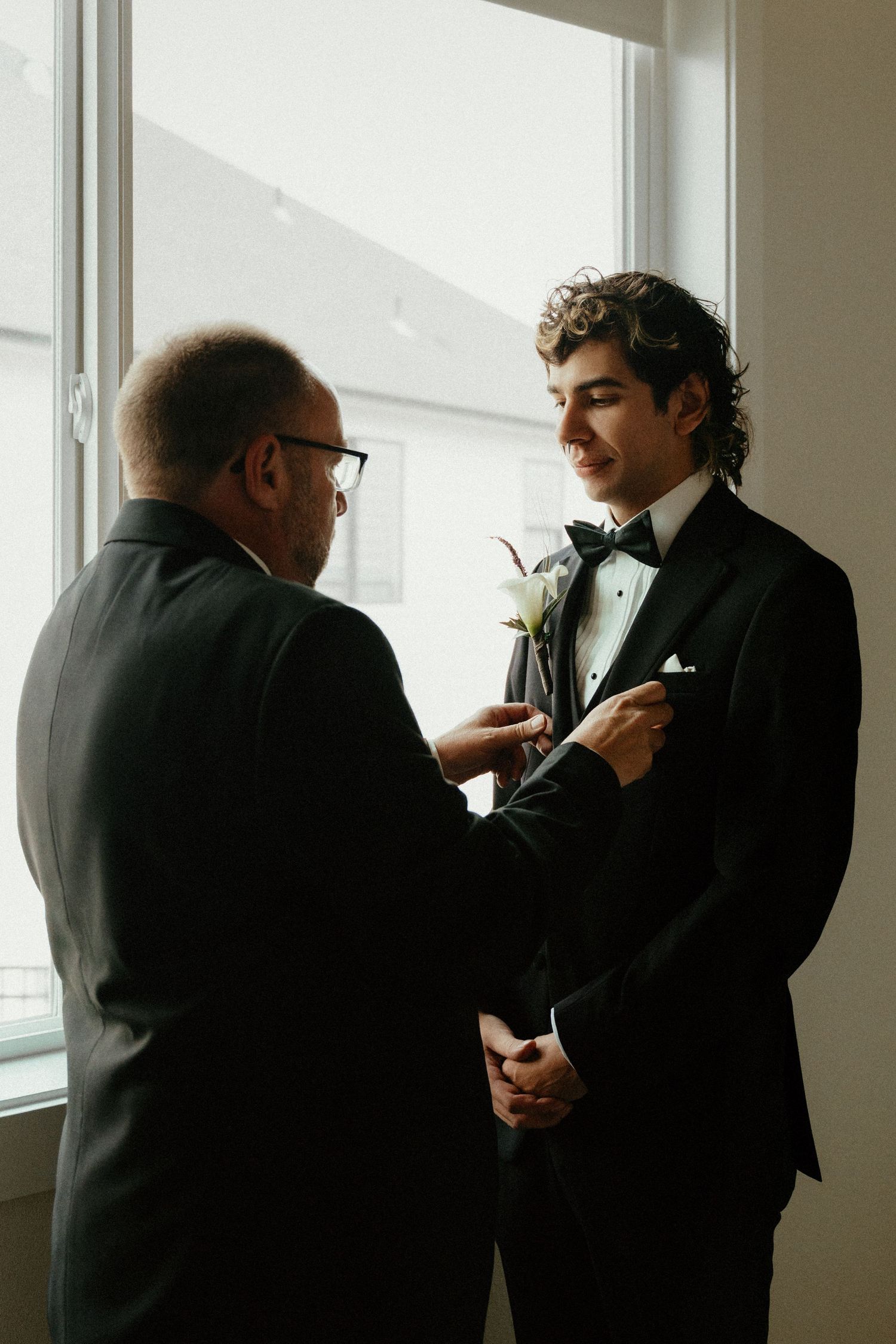 A groom in a black tuxedo has a white boutonniere pinned to his lapel by an older man near a bright window.