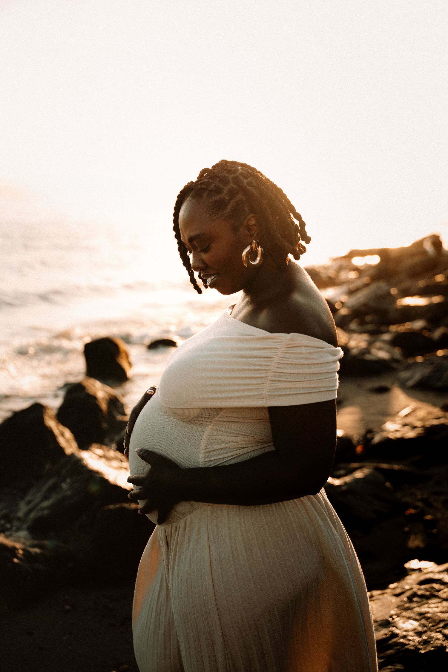 Pregnant woman in white off-shoulder gown poses on rocky beach at golden hour sunset.