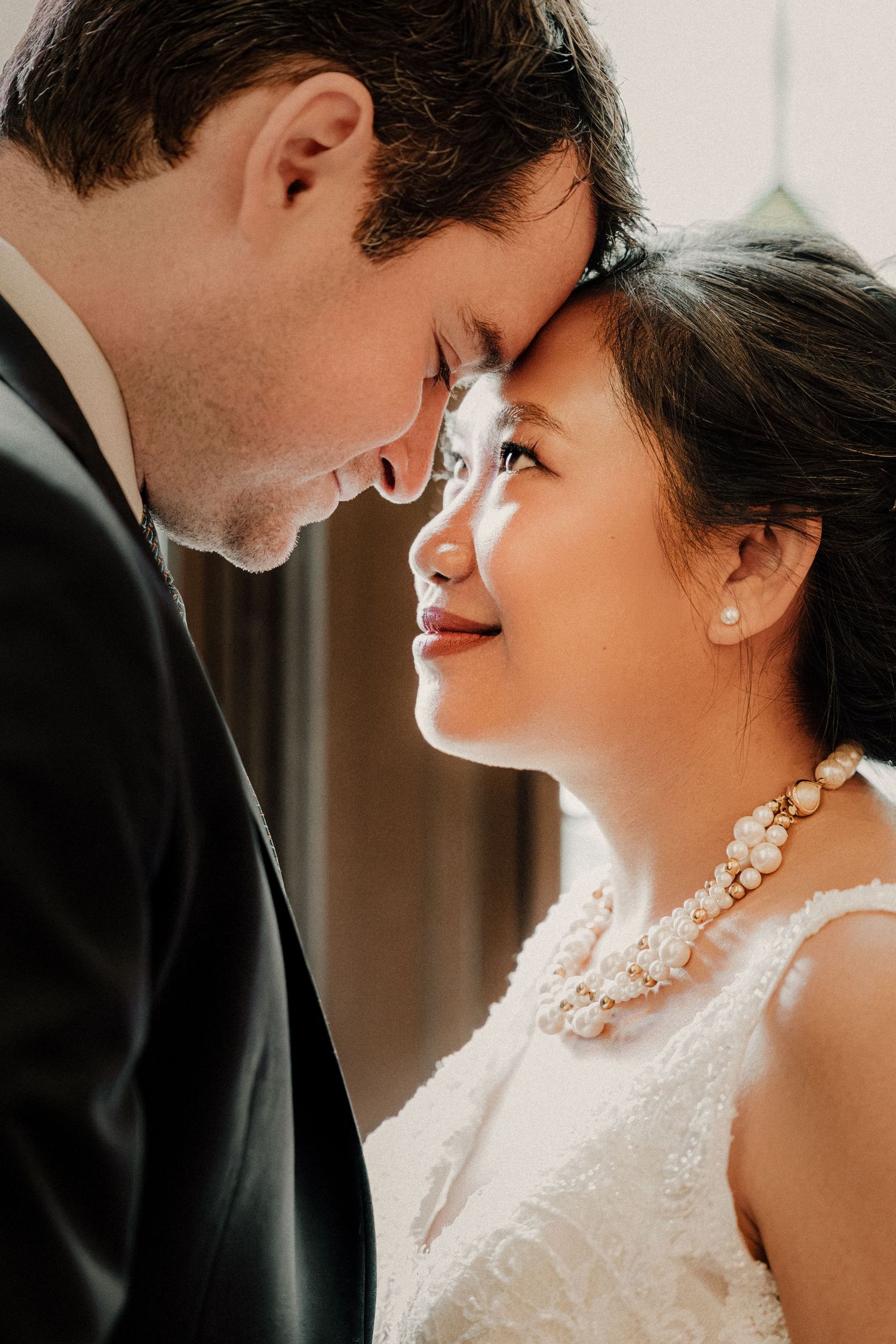 A bride and groom touch foreheads tenderly in an intimate wedding portrait, she wearing a pearl necklace and white lace gown.