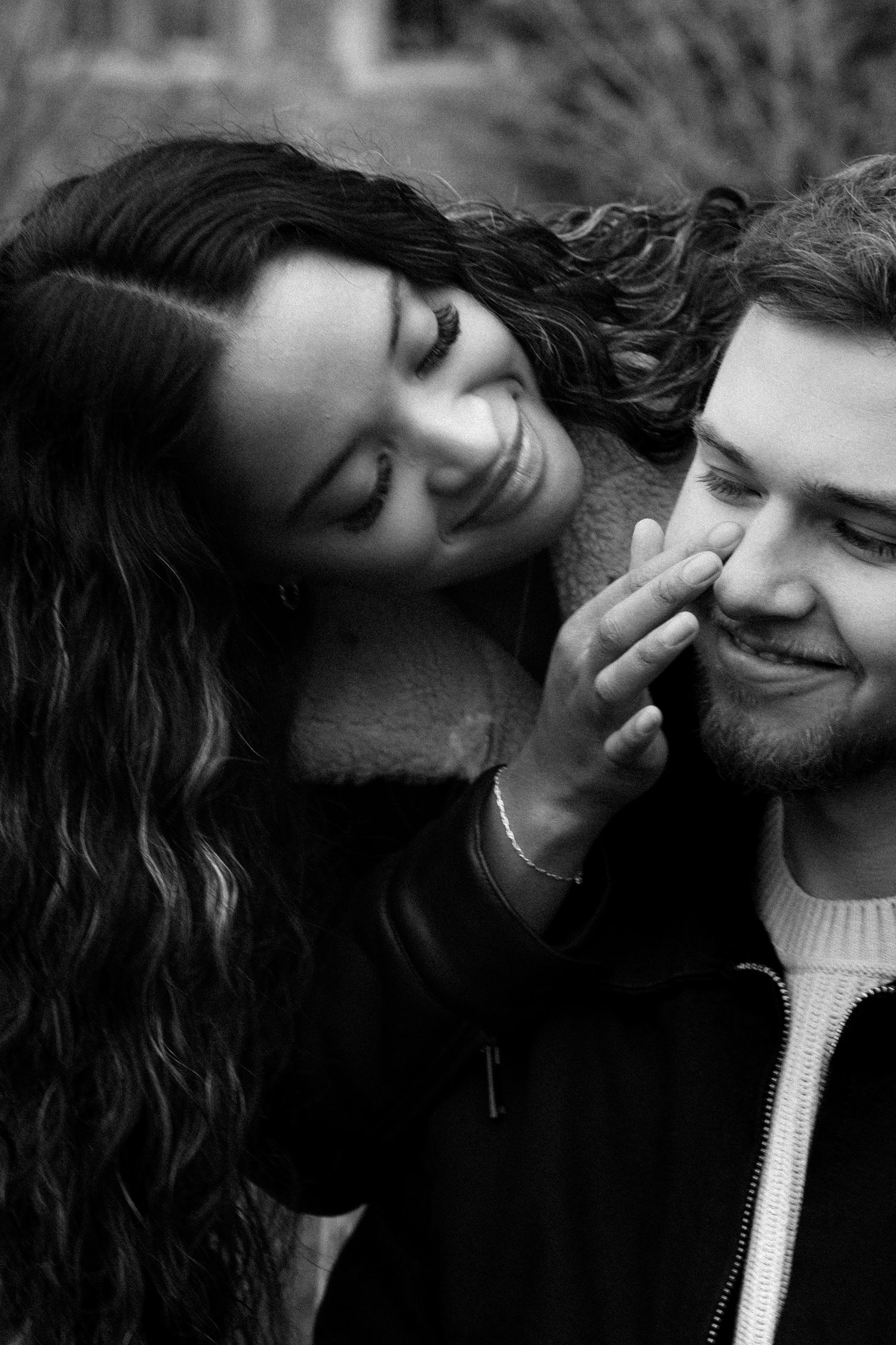 Black and white photo of a smiling couple, woman tenderly touching mans cheek, sharing a romantic moment outdoors.