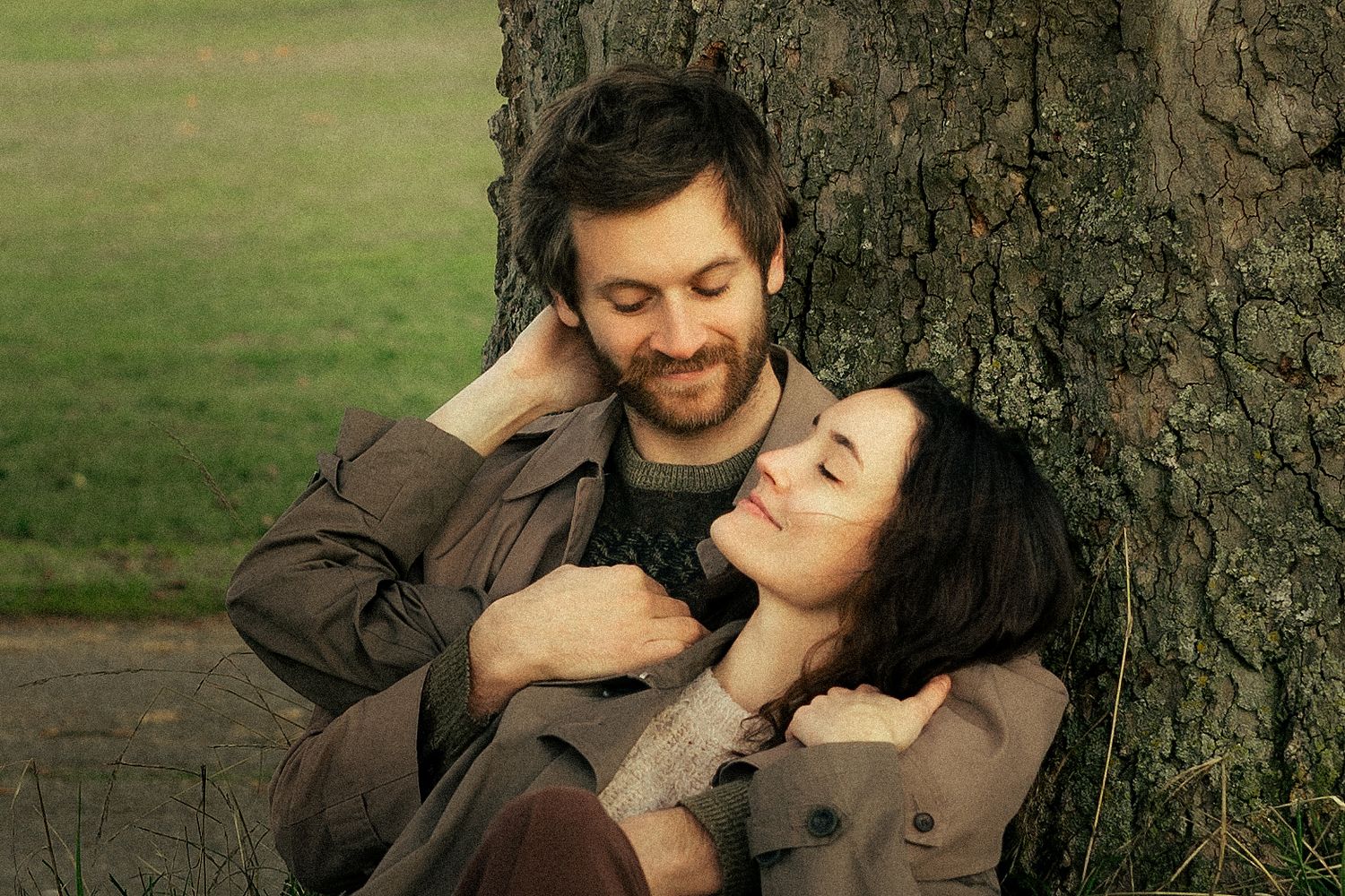 A couple sits together against a tree in a park, smiling and embracing in a romantic outdoor moment.