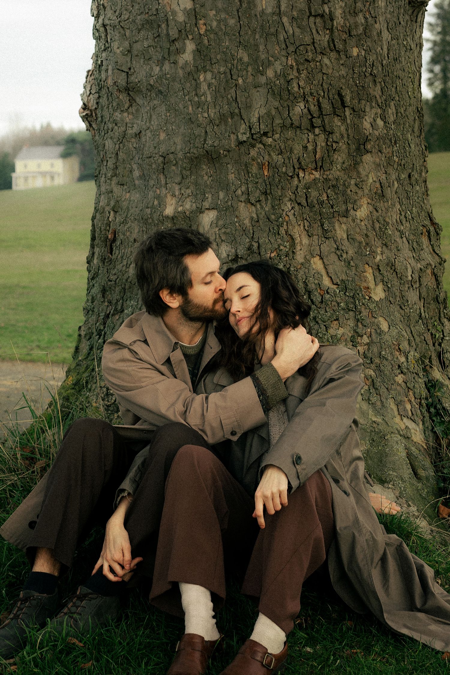 A couple sits leaning against a tree trunk in a park, sharing a tender kiss while dressed in earthy toned coats.
