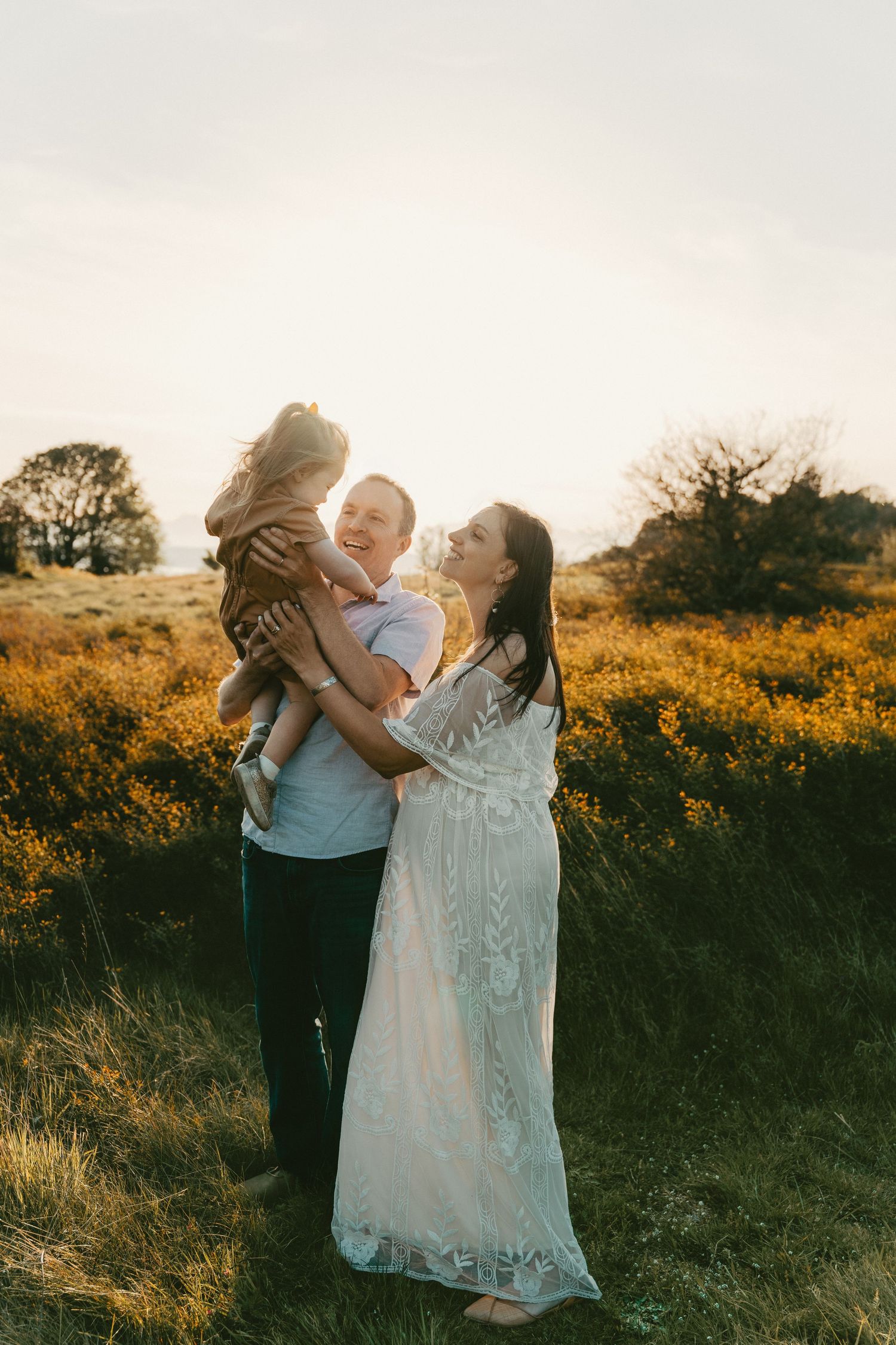 A happy family of three poses in a golden field at sunset, with parents smiling at their young child.