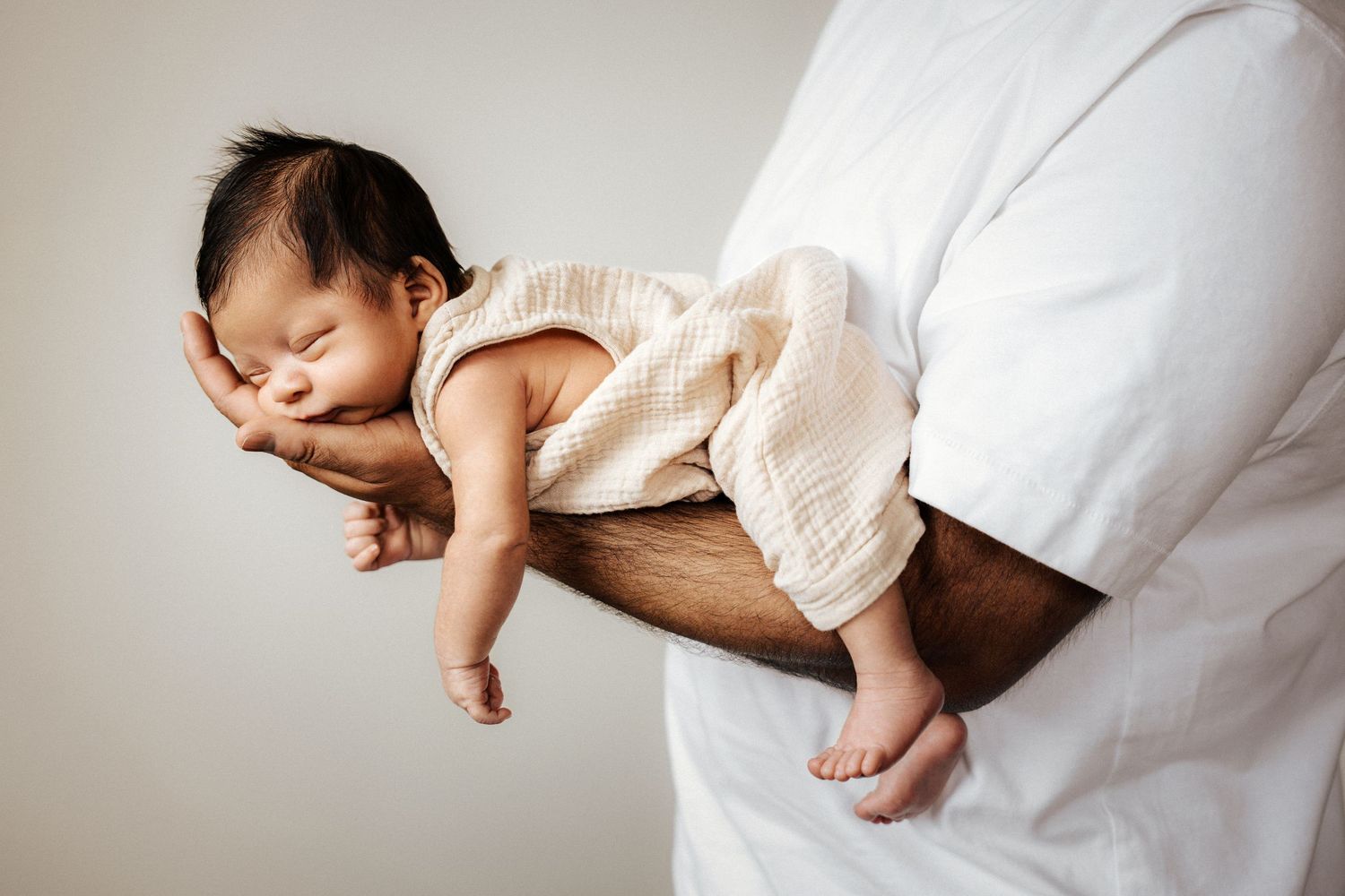 Newborn baby sleeping peacefully on father's arm, wearing cream muslin romper against white background.