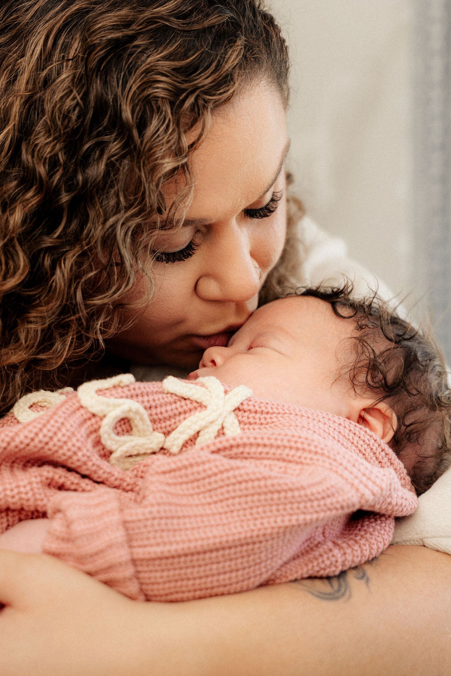 A mother with curly hair tenderly kisses her sleeping newborn baby wrapped in a pink knit outfit.