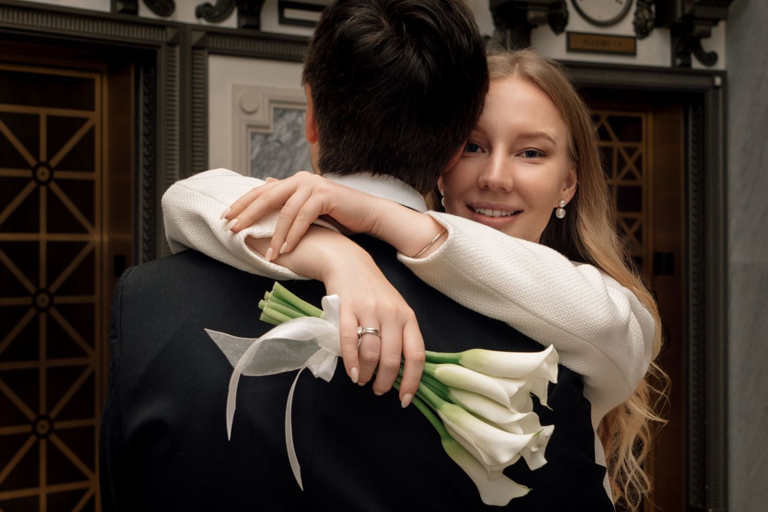 Bride in white dress hugging groom, holding white calla lily bouquet, showing engagement ring, smiling over his shoulder.