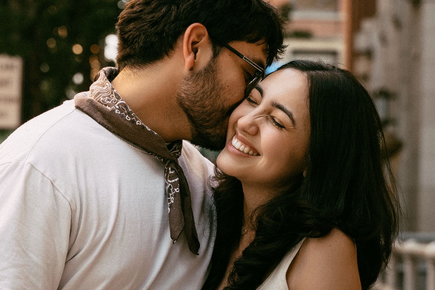 A happy couple sharing a tender moment, man kissing smiling woman wearing glasses outdoors.