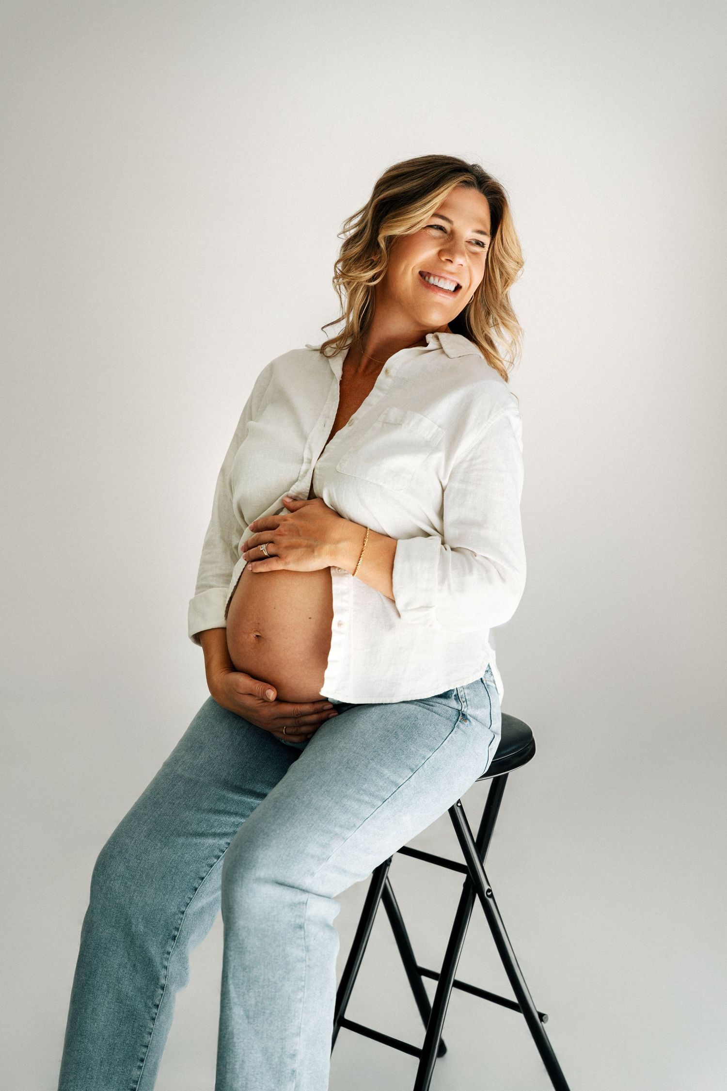 Smiling pregnant woman in white shirt and jeans sitting on stool, cradling her baby bump against white background.