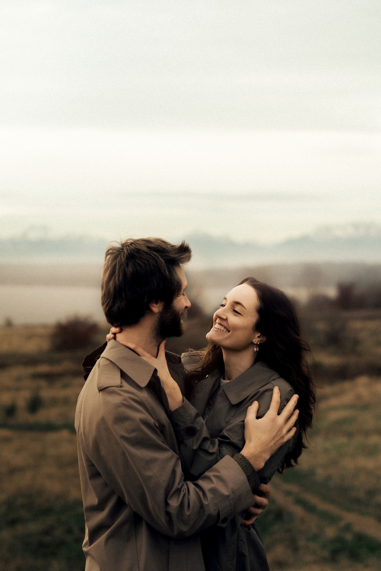 A smiling couple embraces face-to-face in an open field with mountains visible in the distance.