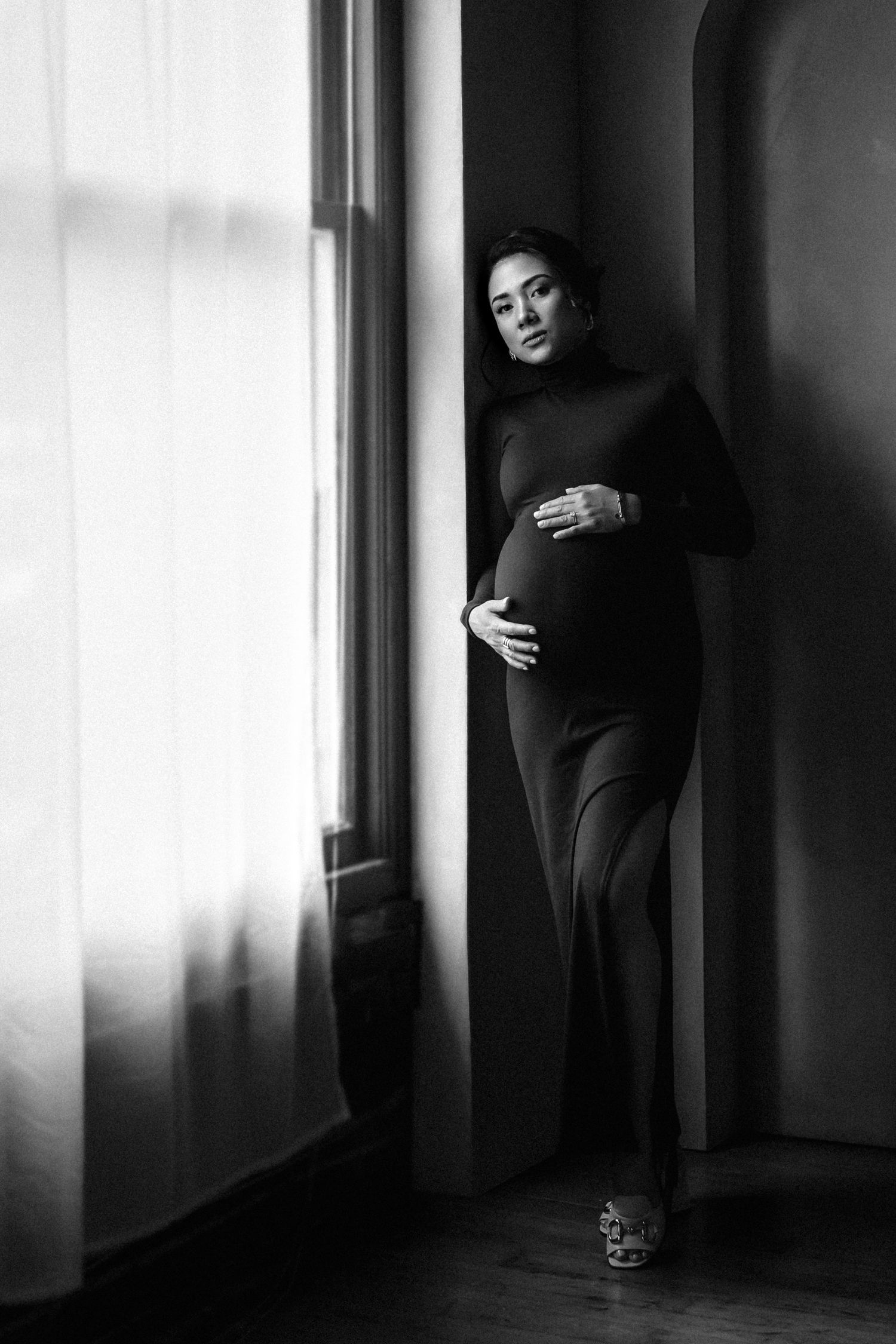 Black and white portrait of a pregnant woman in a dark dress standing by a window indoors.