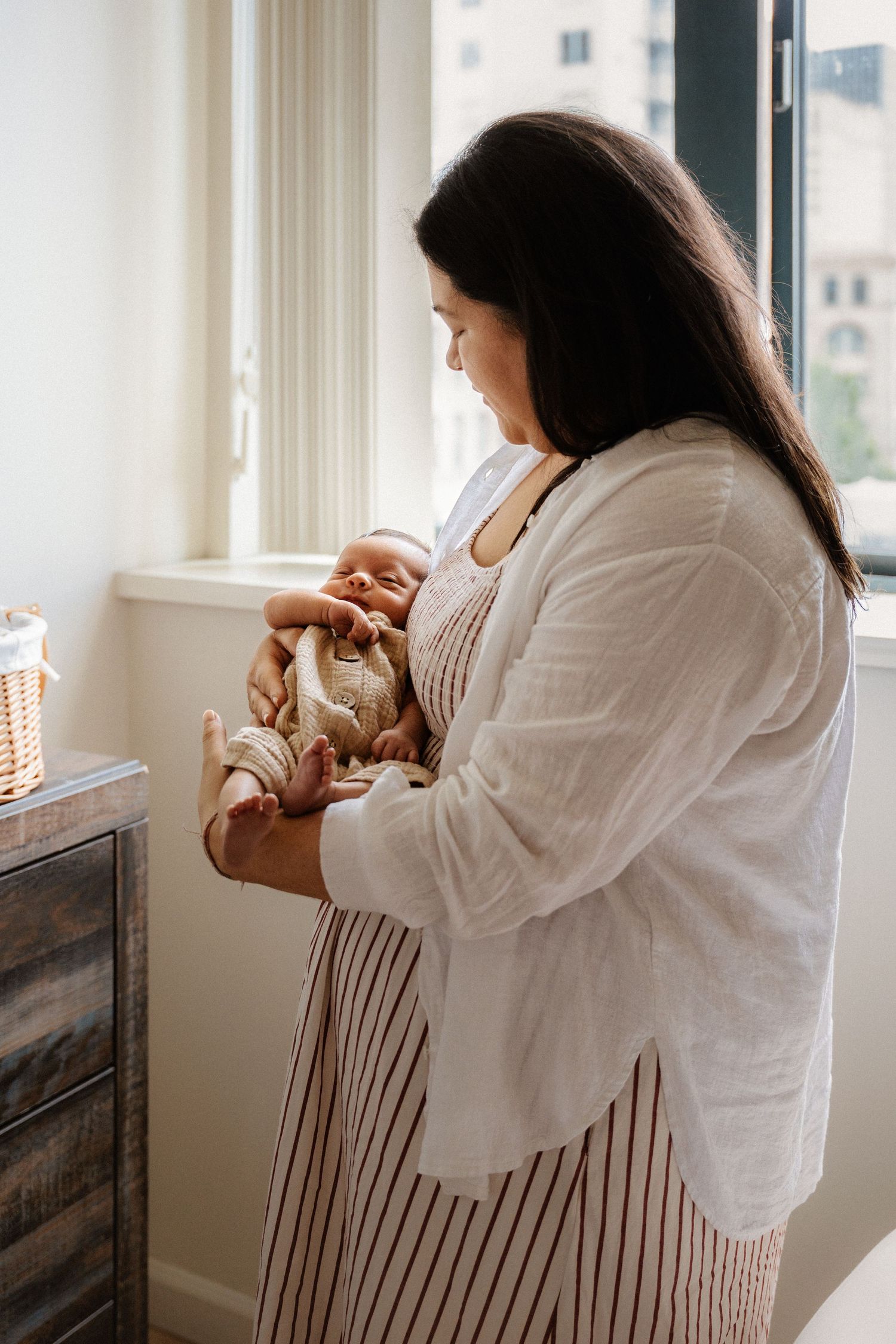 A mother with dark hair holds her sleeping newborn baby near a bright window, wearing a striped dress and white shirt.