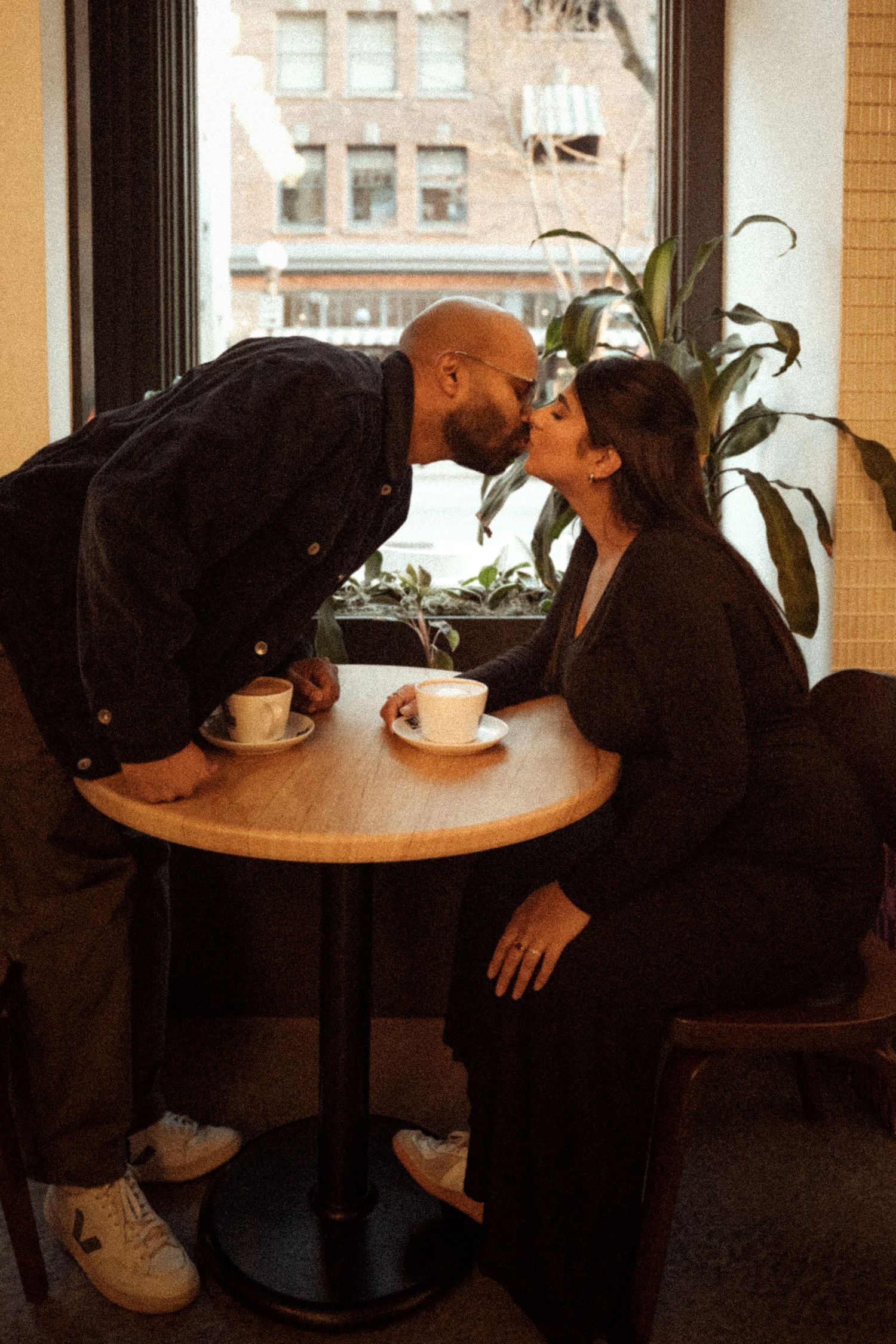 A couple shares a romantic kiss over coffee at a cozy cafe table with a city view.