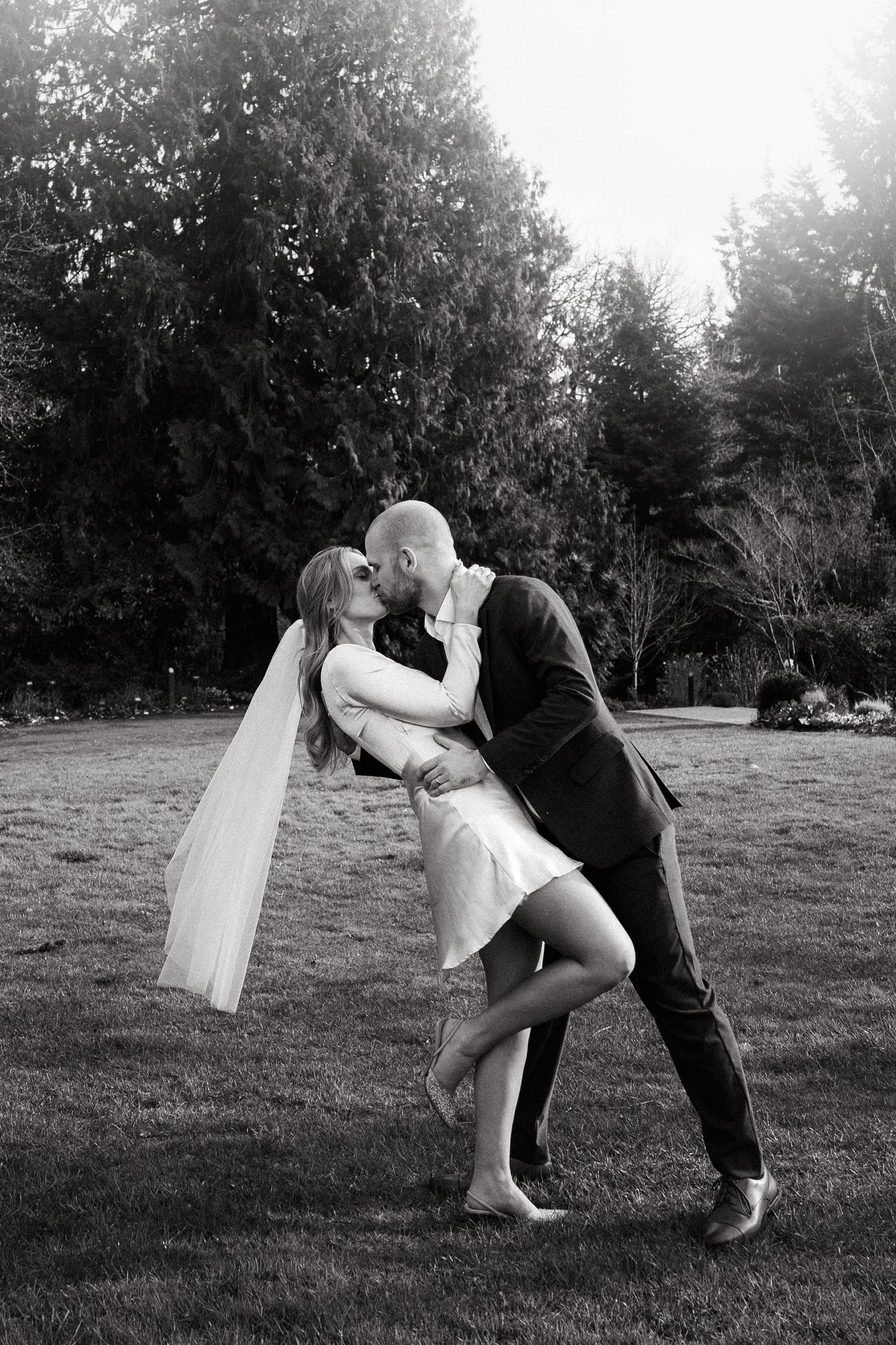 Black and white photo of bride and groom sharing a romantic dip kiss outdoors on lush green lawn with trees.