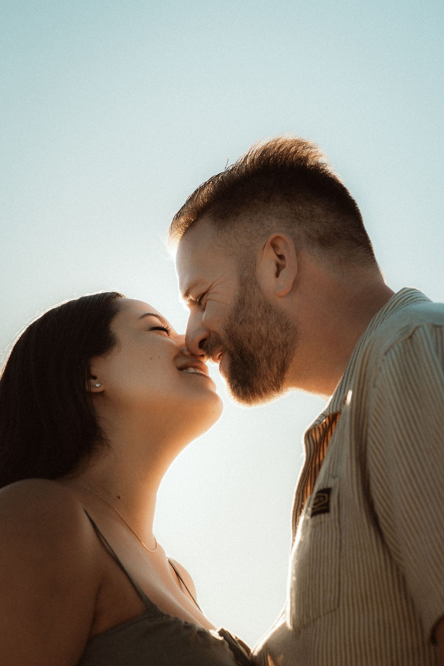 A couple shares a romantic almost-kiss backlit by a clear blue sky in soft sunlight.