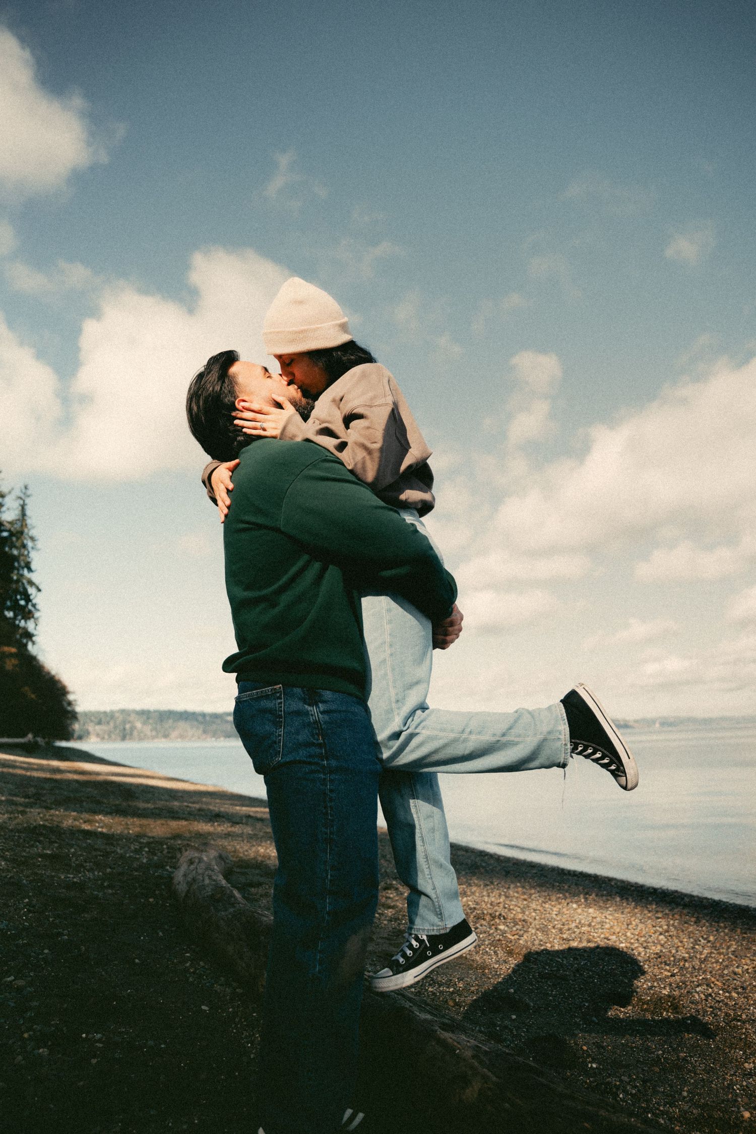 Romantic couple kissing on a rocky beach shore, man lifting woman under a blue cloudy sky.