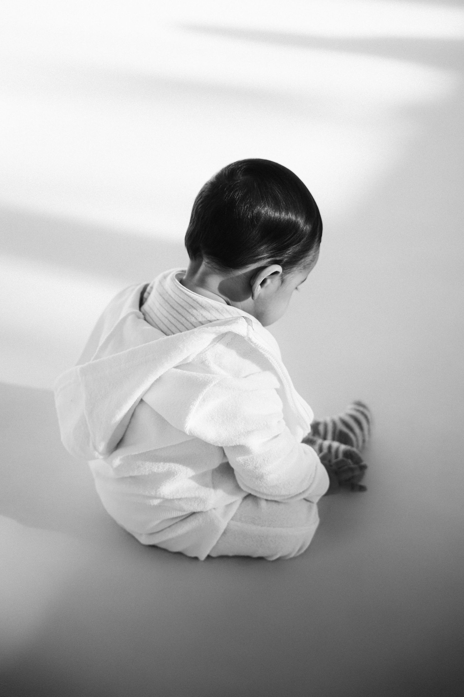 Black and white photo of a young child sitting on floor with back turned, wearing white outfit and striped socks.