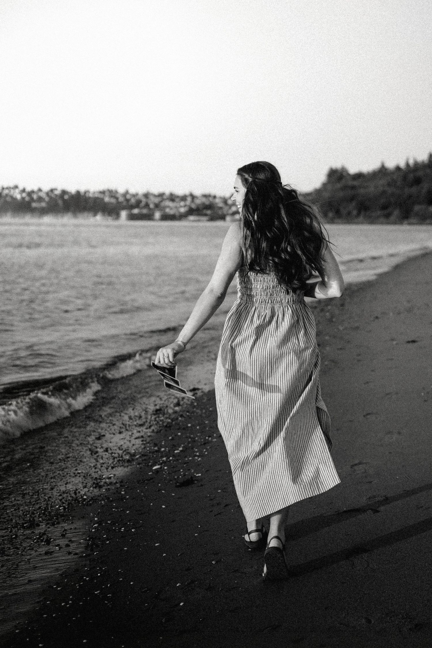 Woman in long dress walking barefoot along beach shoreline, carrying shoes, with ocean waves and trees in background.