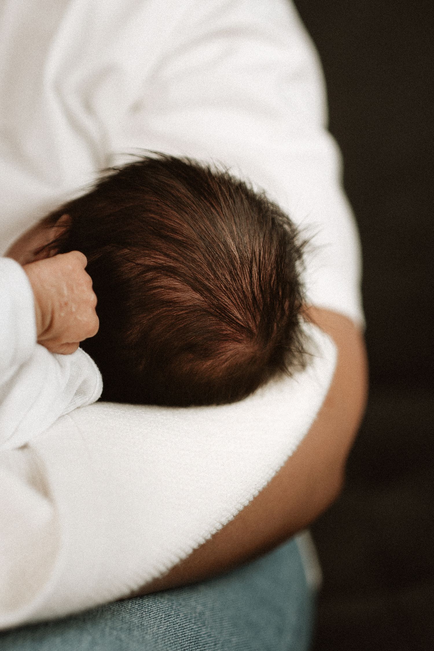 A newborn baby with dark hair being cradled in the arms of a person wearing a white sweater and blue jeans.