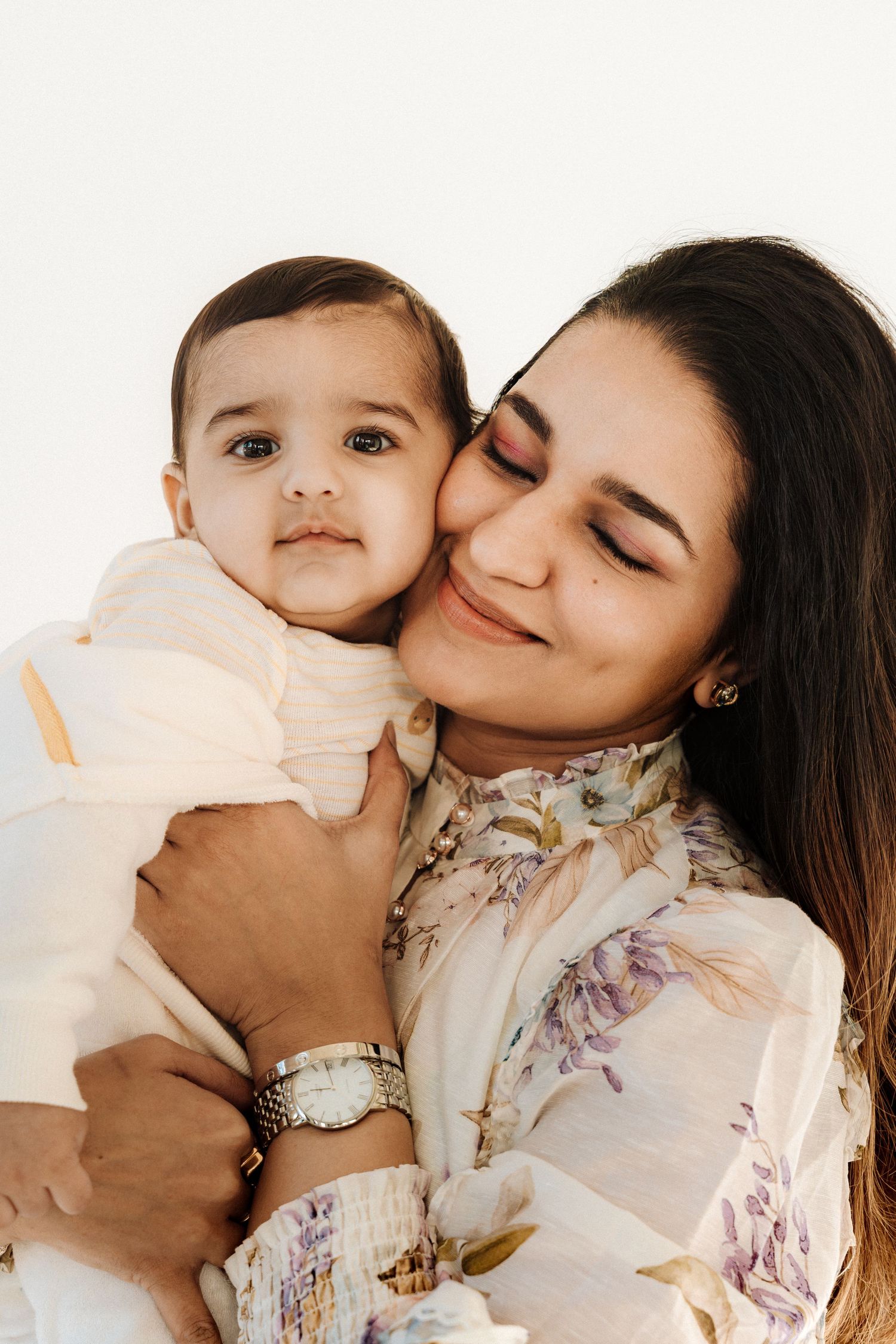 A smiling mother tenderly holds her baby close, both wearing soft floral outfits against a white background.