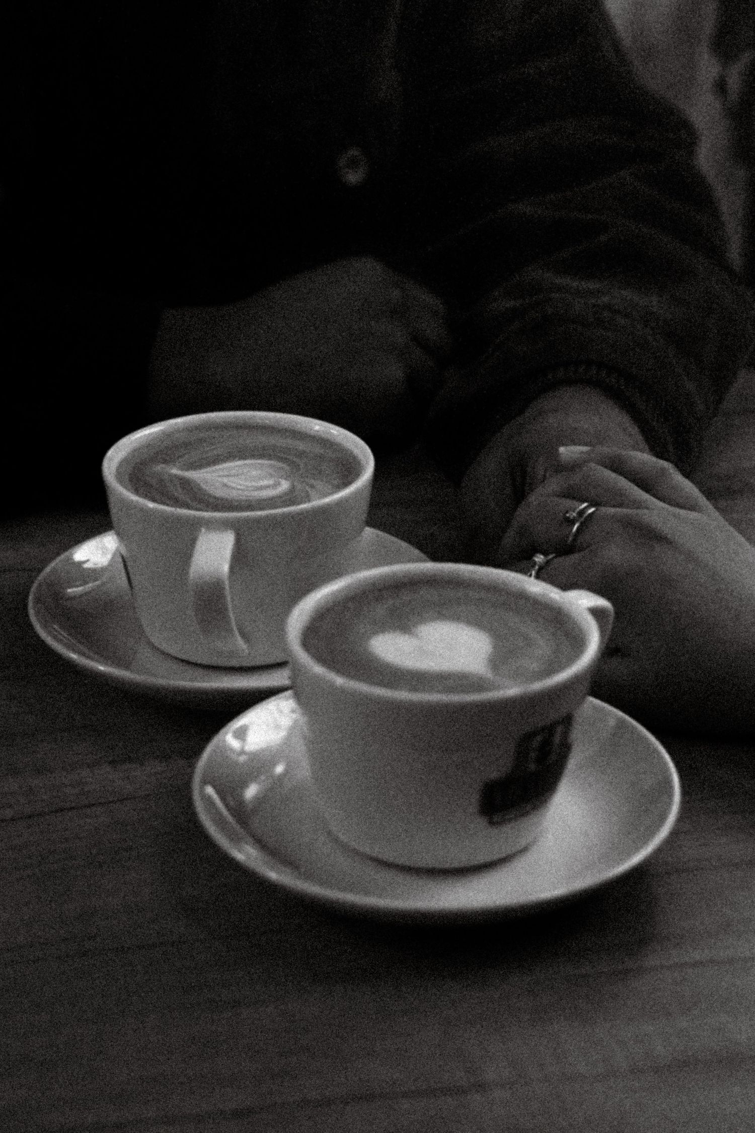 Black and white photo of two latte art coffees on saucers with hands visible on a dark wooden table.