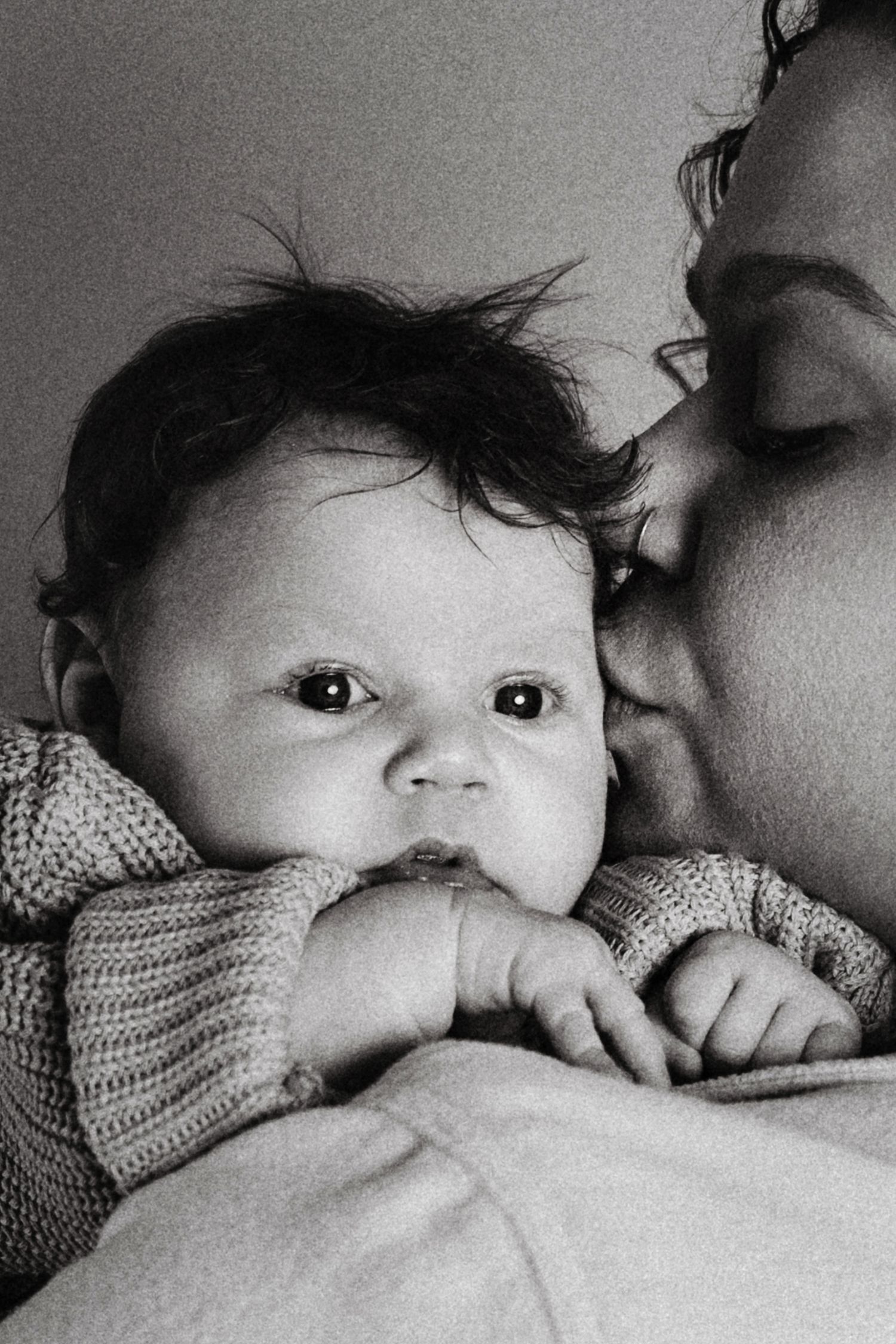 Black and white photo of a newborn baby with dark hair being kissed by mother, looking at camera.