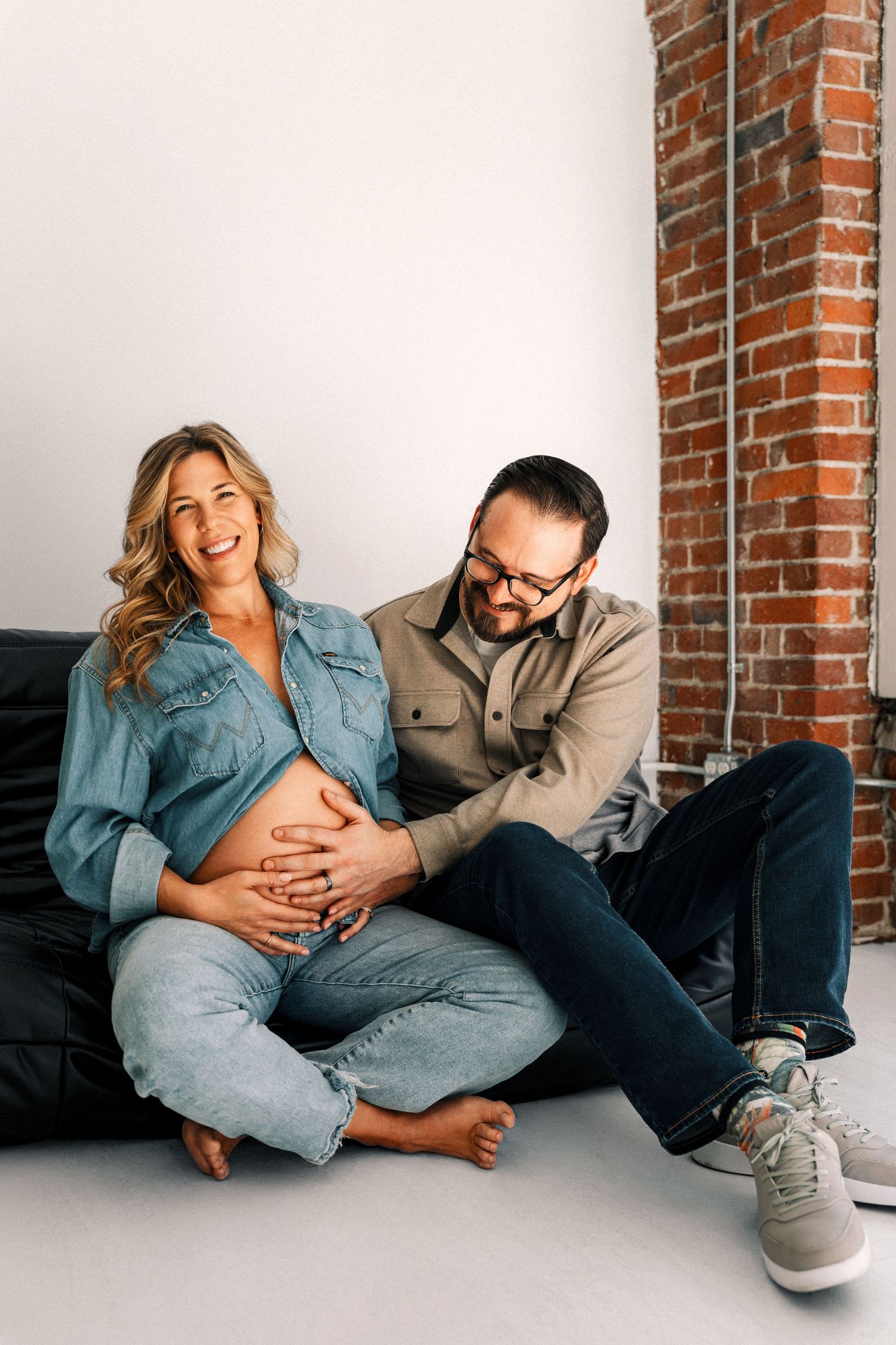 Happy pregnant woman in denim and man with glasses sitting on black couch, both touching baby bump.