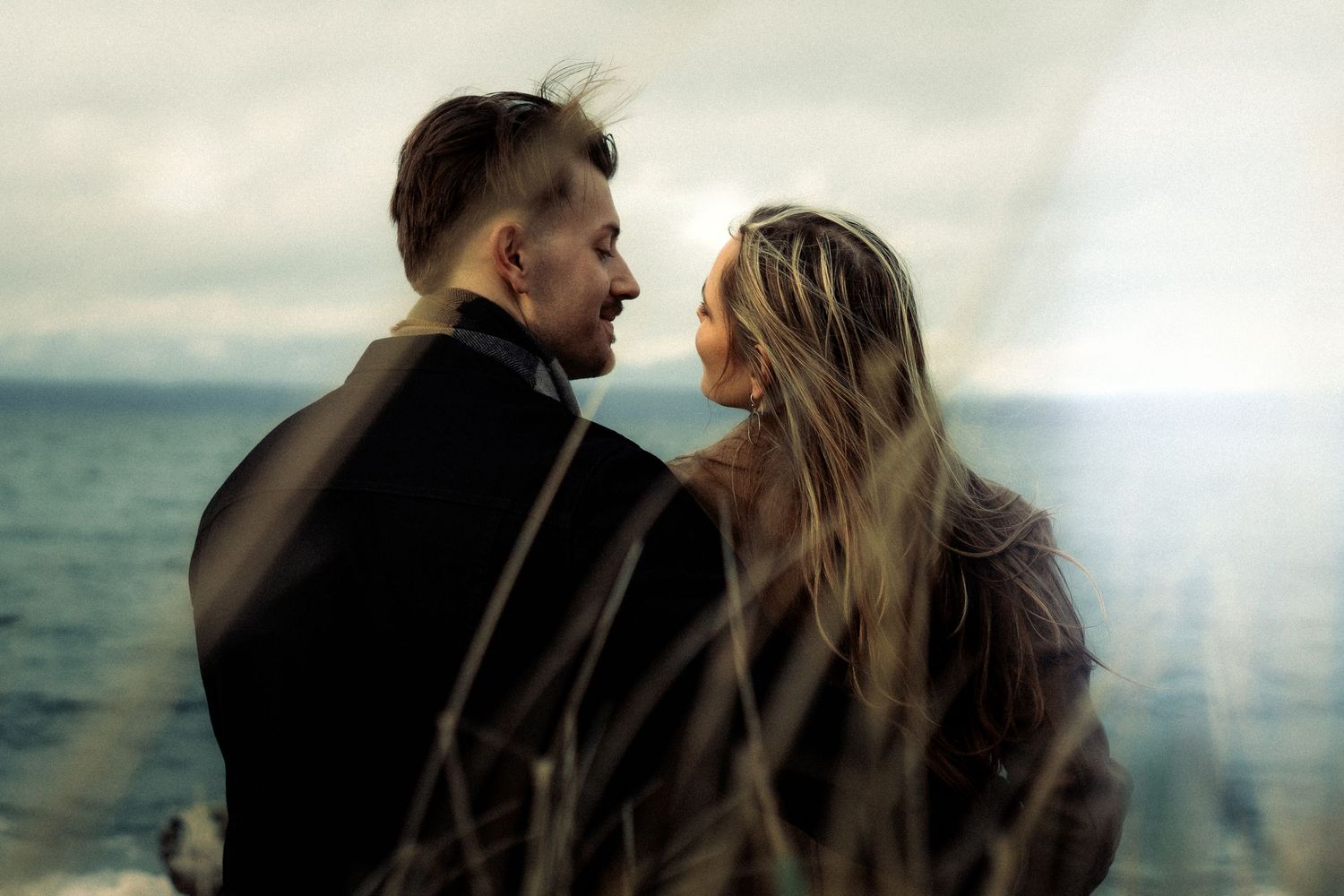 Romantic couple sharing an intimate moment by the sea, with wind-blown hair against a dramatic cloudy sky.