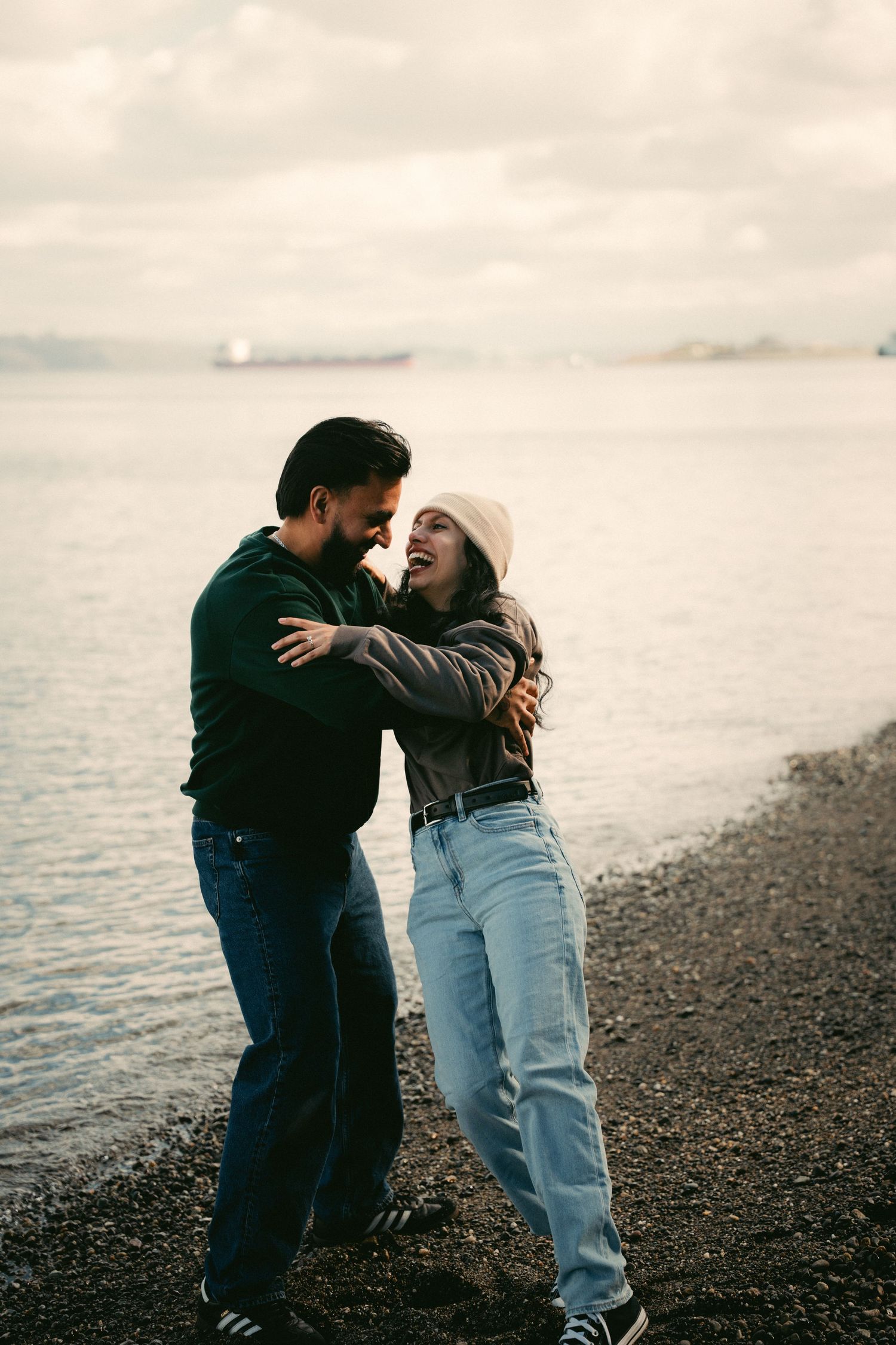 Couple laughing and embracing on a rocky beach shoreline, wearing casual winter clothes with ocean view behind them.