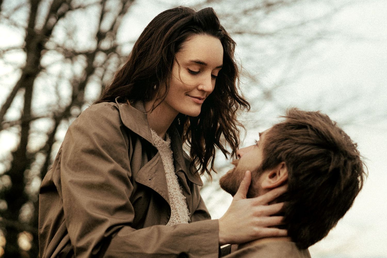 A romantic couple sharing a tender moment outdoors, woman smiling down at man while holding his face gently.