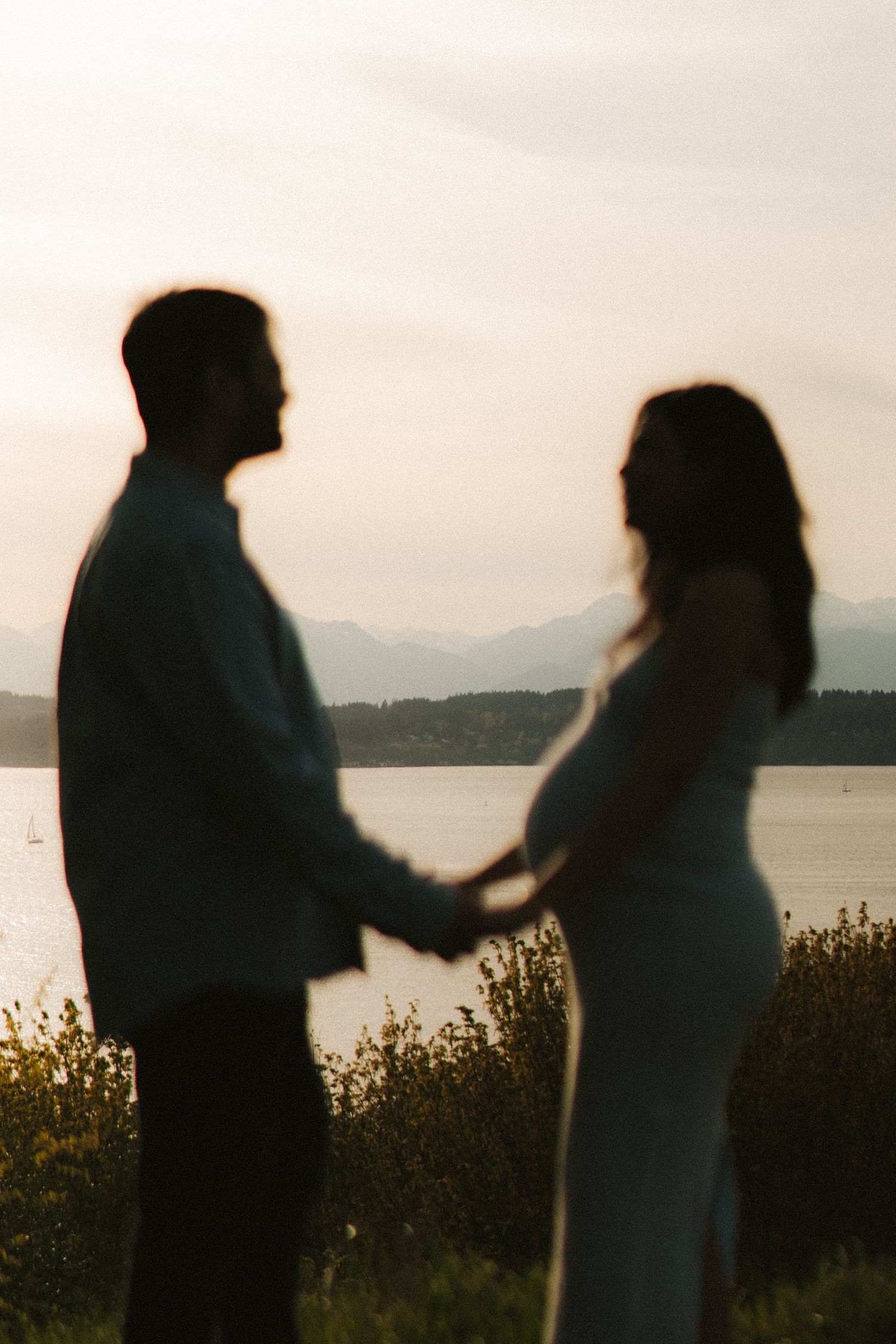 Silhouette of a couple holding hands near a lake with mountains at sunset, woman showing a baby bump.
