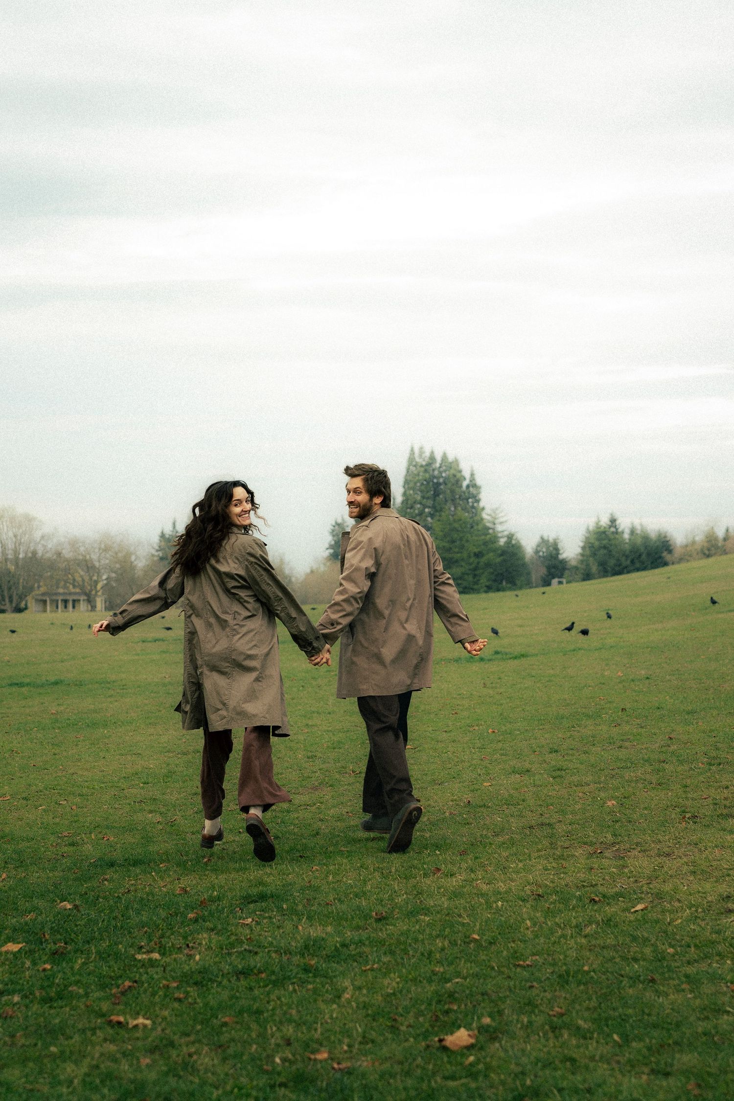 A joyful couple in matching trench coats holds hands while running through a lush green park on a cloudy day.